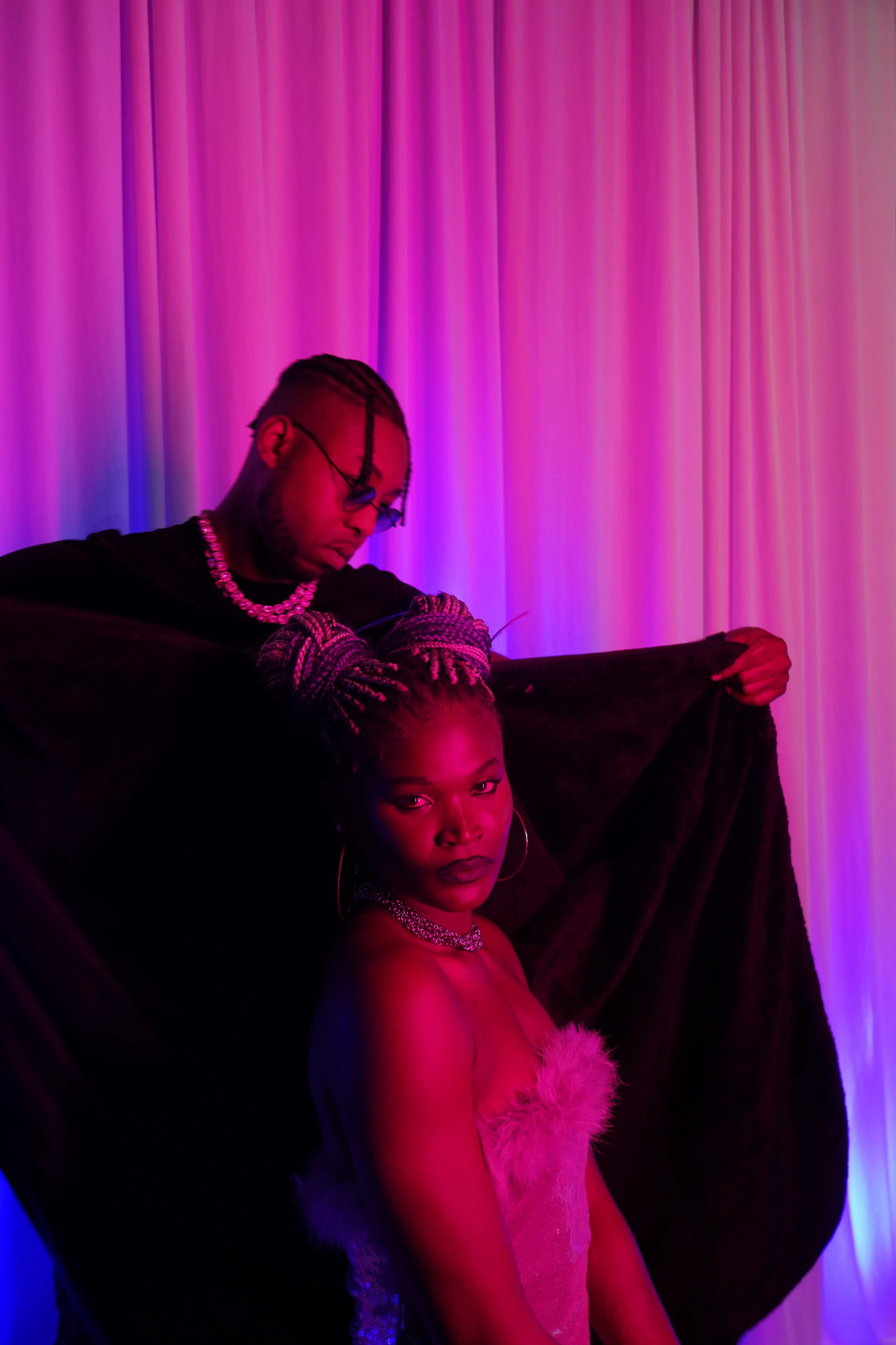 Alpha The Wolf holding the black robe stretched wide behind a woman in a pink feathered dress so the fabric spreads like a pair of dark wings, both staring down the lens under magenta light.