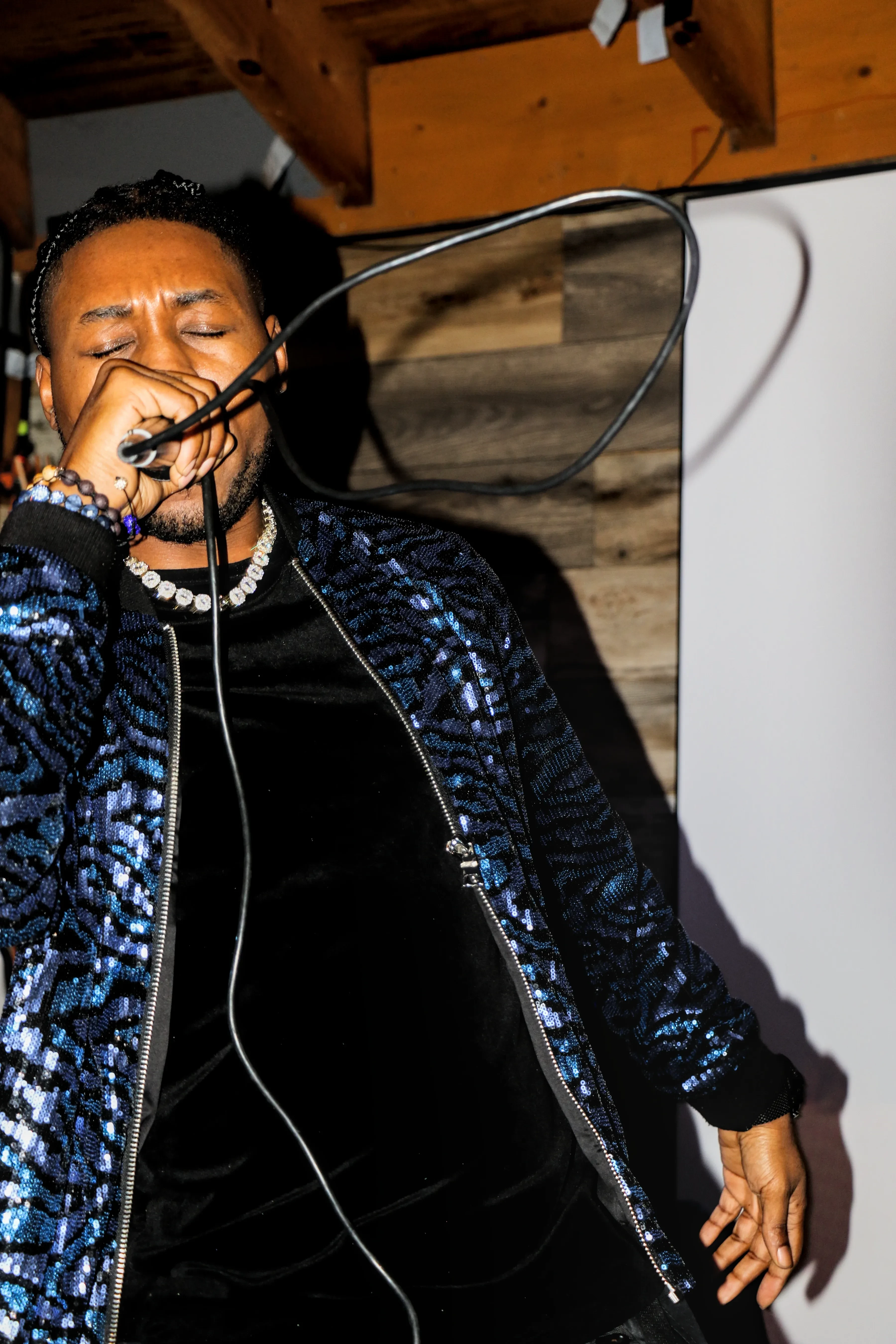 Alpha The Wolf close in the frame with his eyes shut and his fist clamped around the microphone, exposed wooden beams and a reclaimed timber wall behind his shoulder.