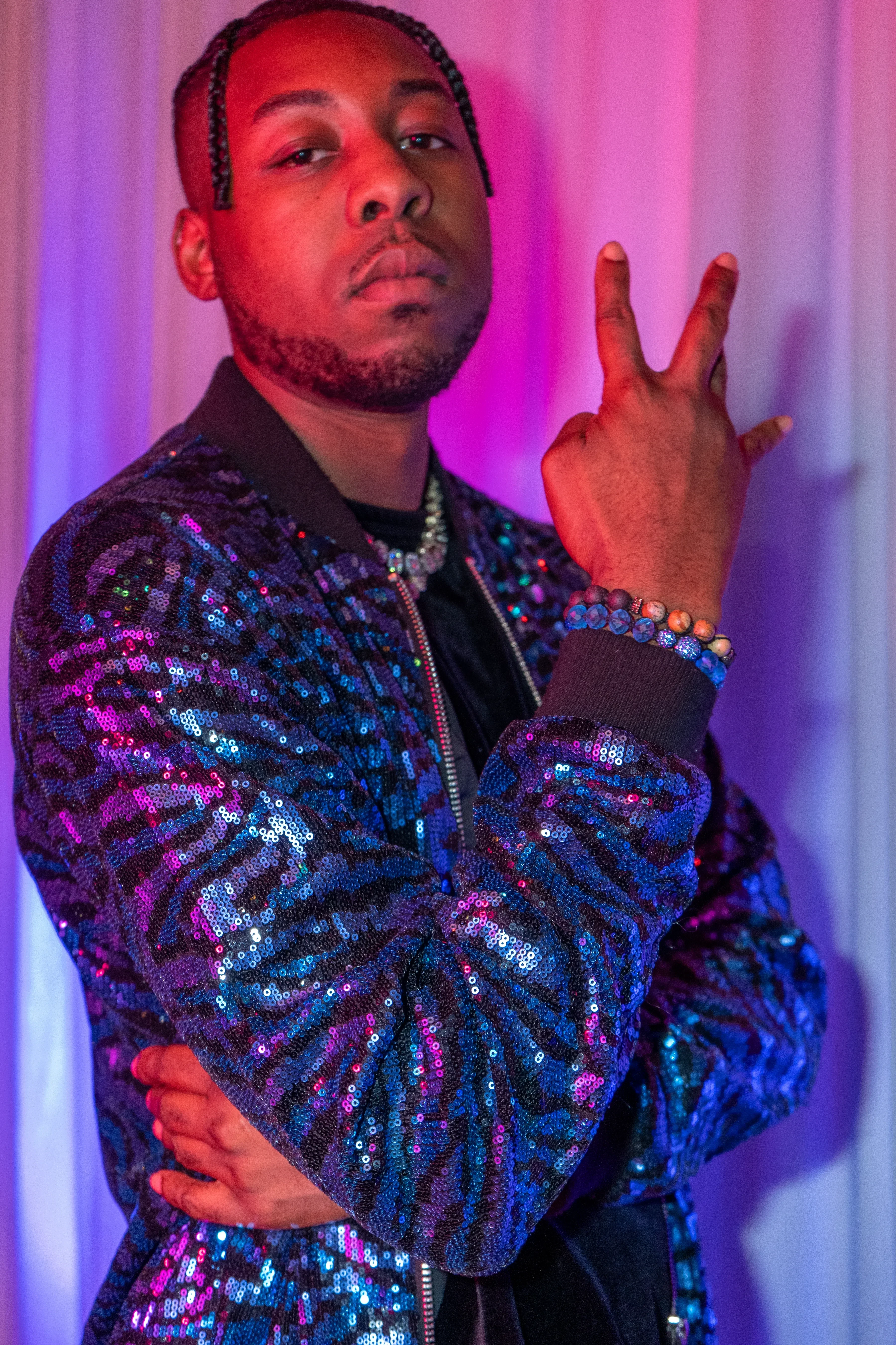Alpha The Wolf in a vertical portrait against the curtain with one hand raised beside his face, fingers parted, arms crossed over the sequinned jacket, his chin lifted and the drapery glowing pink and blue behind