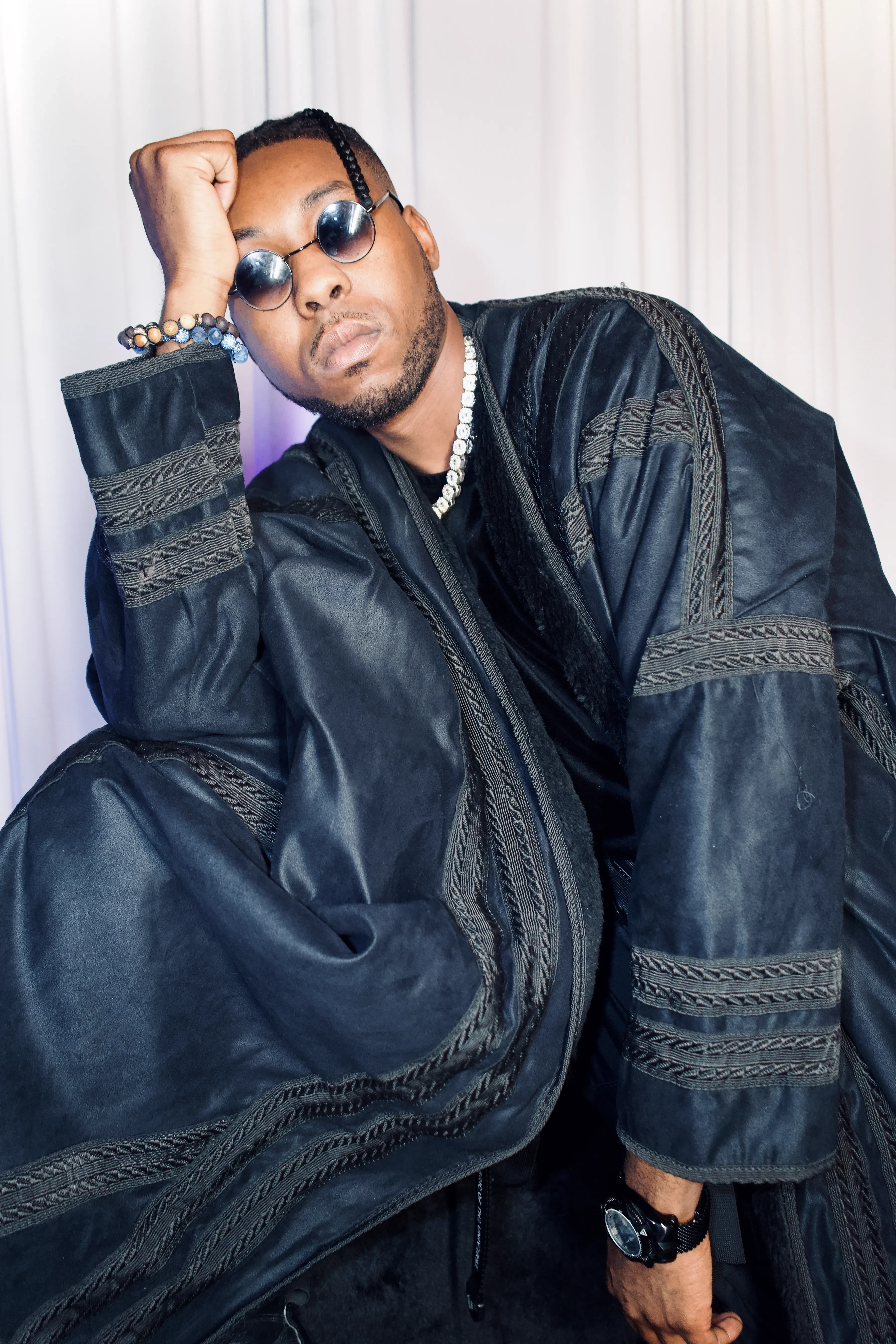 Alpha The Wolf in a full black embroidered robe with an elbow lifted and his hand at his head, round dark sunglasses and a silver chain, the heavy braided trim catching the light against a pale wall.