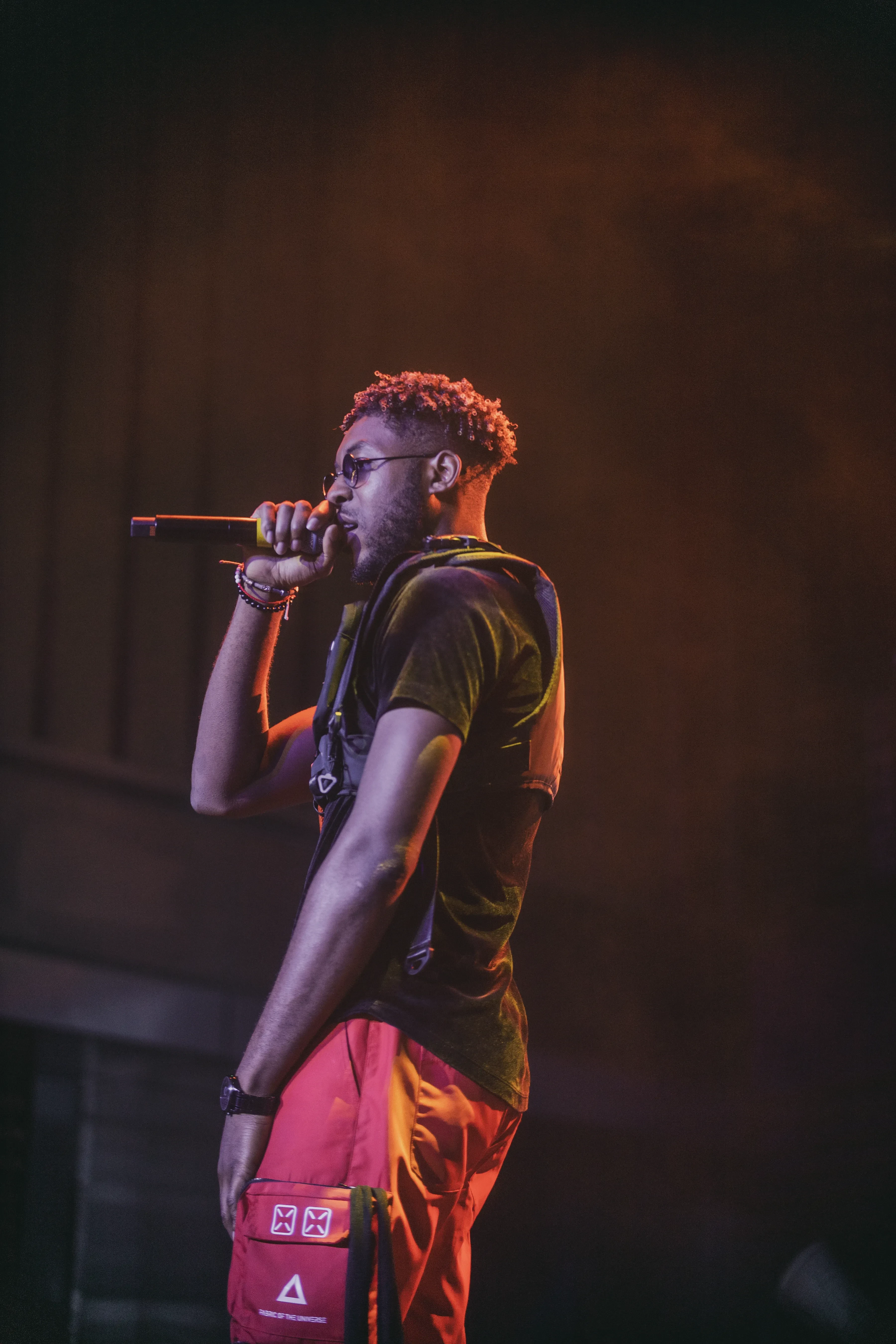 Alpha The Wolf turned nearly away from the camera in profile, mic pressed to his mouth and his other arm hanging loose with a black watch on the wrist, olive-cast black tee, red cargo pants with a triangle logo on the pocket, deep orange haze filling the frame