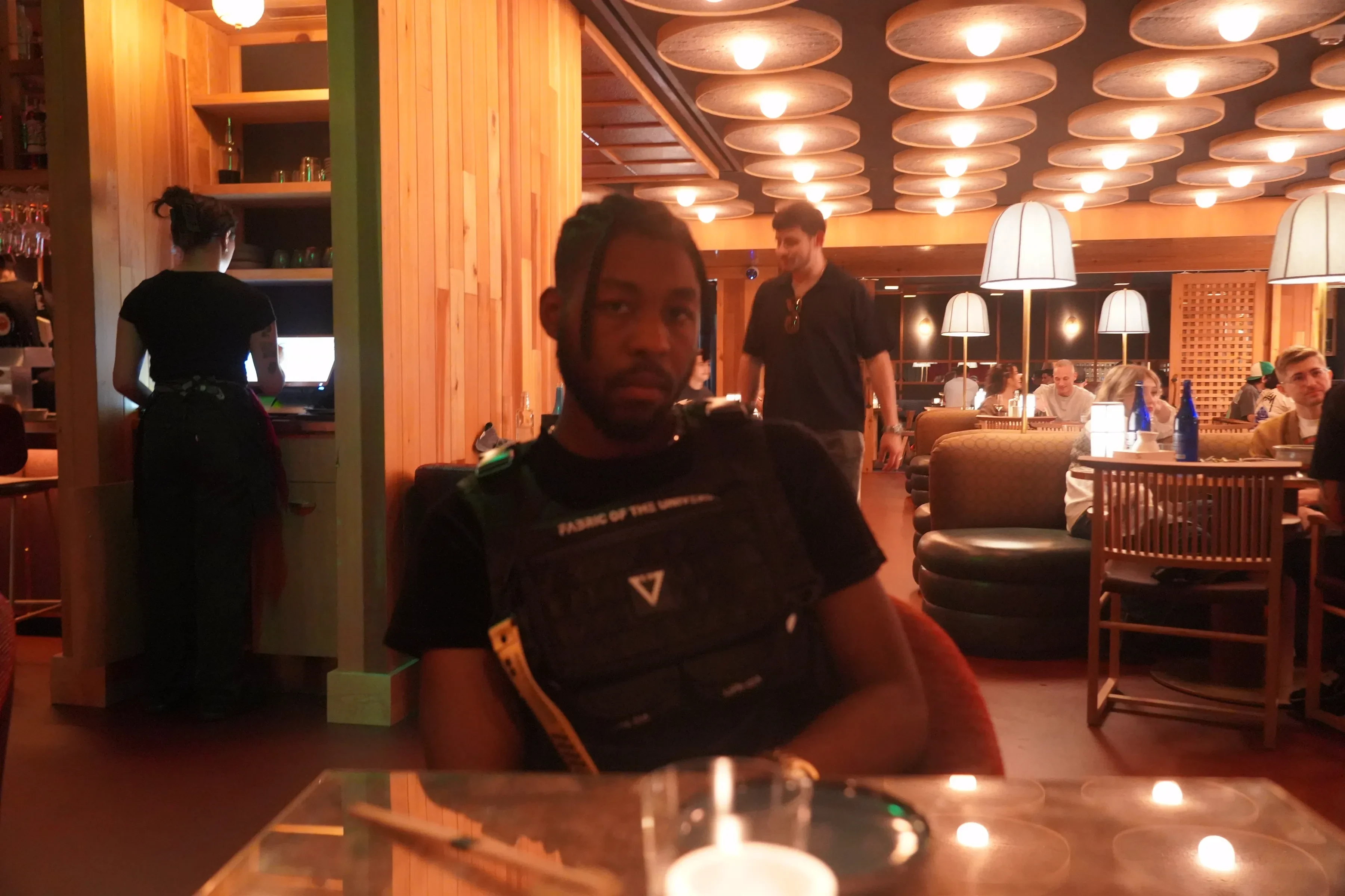 Alpha The Wolf looking directly into the lens across the table, the harness straps and its yellow accent cord clearly visible, a man in a dark shirt walking past behind him
