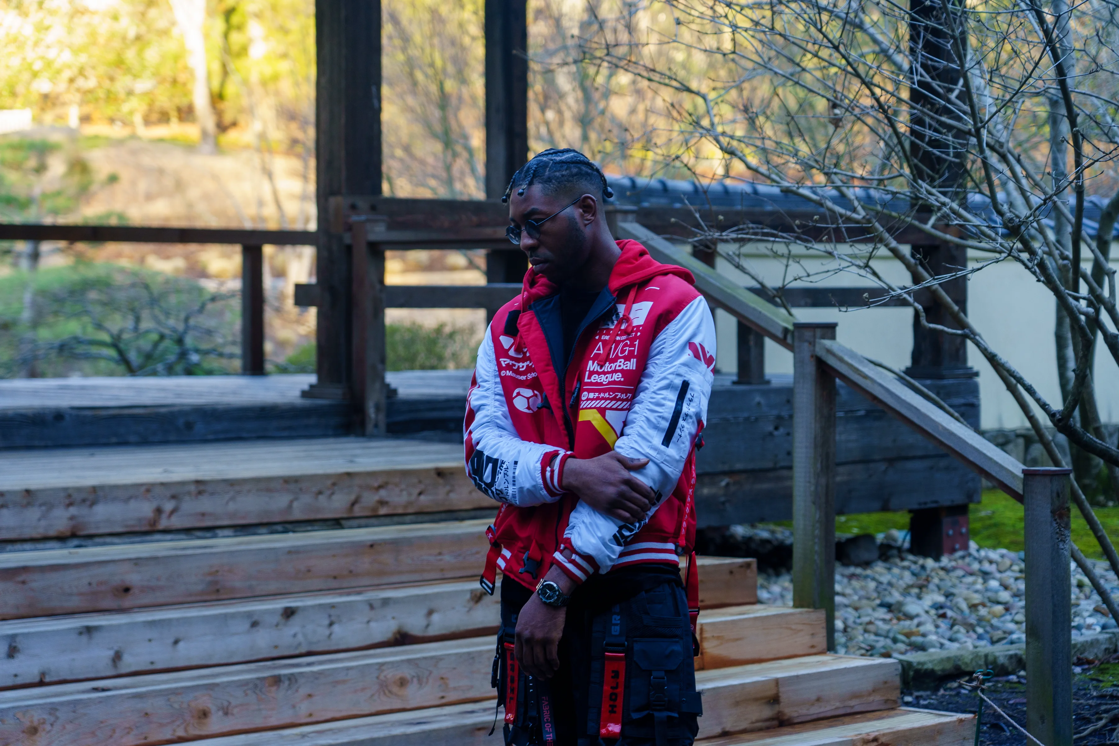 Alpha The Wolf standing at the base of wooden garden steps beside a traditional Japanese pavilion, arms crossed and gaze downward, a bold red and white racing jacket covered in logos and patches, bare winter trees behind him.