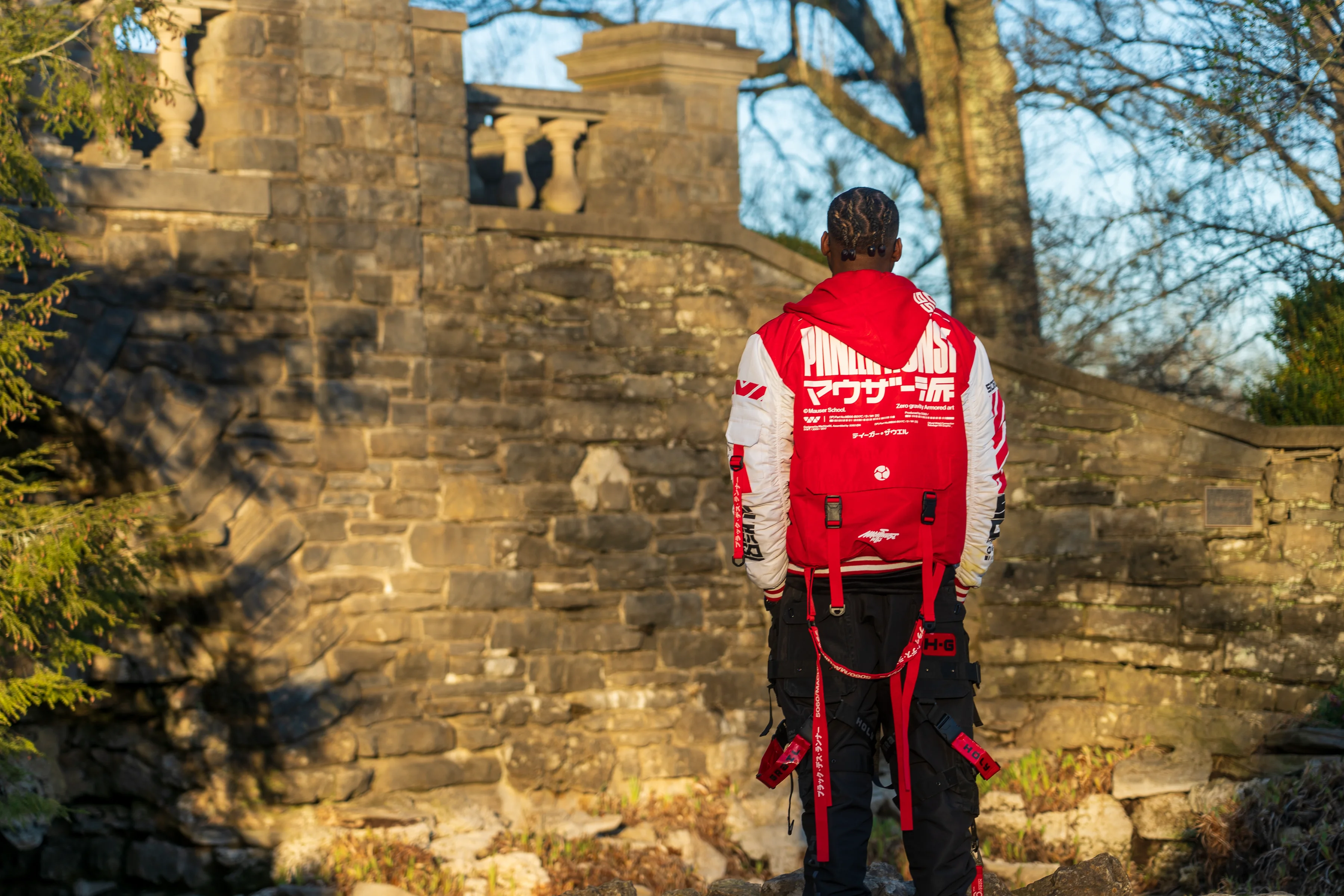 Alpha The Wolf shot from behind before a moss-covered stone archway wall in golden light, the back of his red and white Panzer Kunst racing jacket fully visible, black cargo pants with red utility straps.