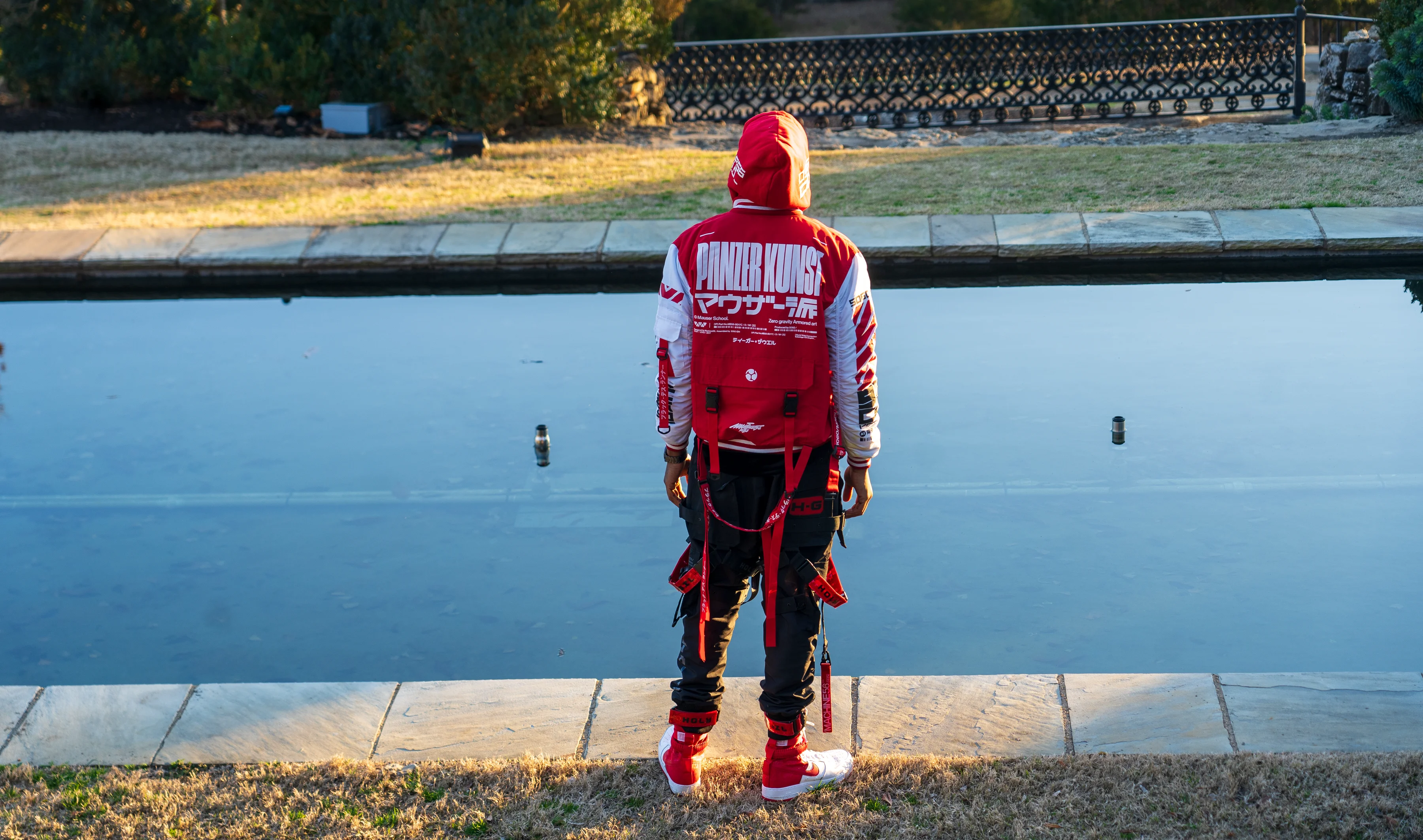 Alpha The Wolf standing with his back to the camera at the edge of a still reflecting pool, the red and white Panzer Kunst jacket reading clearly across his back, black cargo pants with red utility straps.