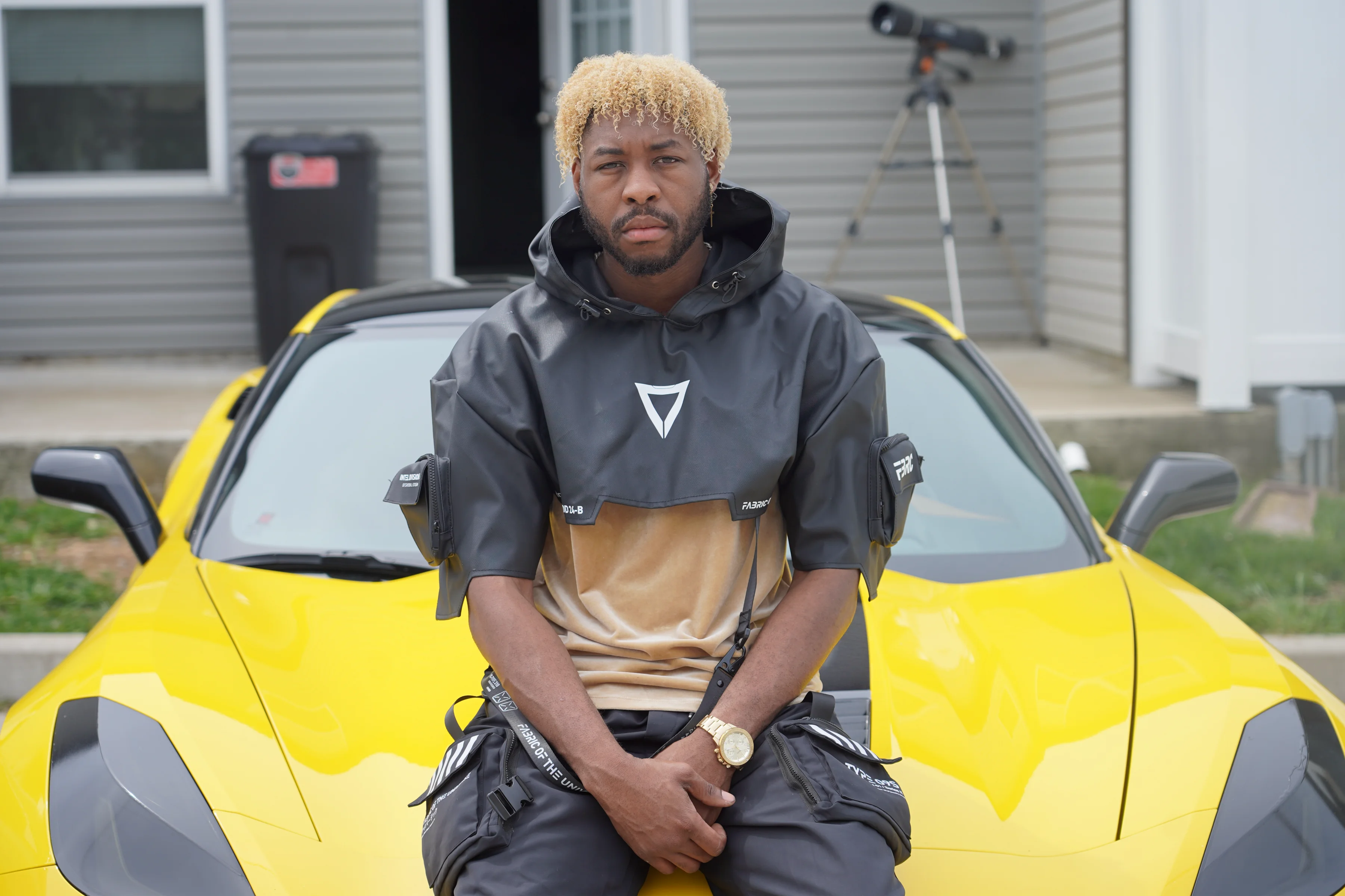 Alpha The Wolf seated on the hood of a yellow Corvette, hands clasped in his lap, staring straight into the camera with a cool, unapologetic expression, a dark grey tactical anorak and gold watch