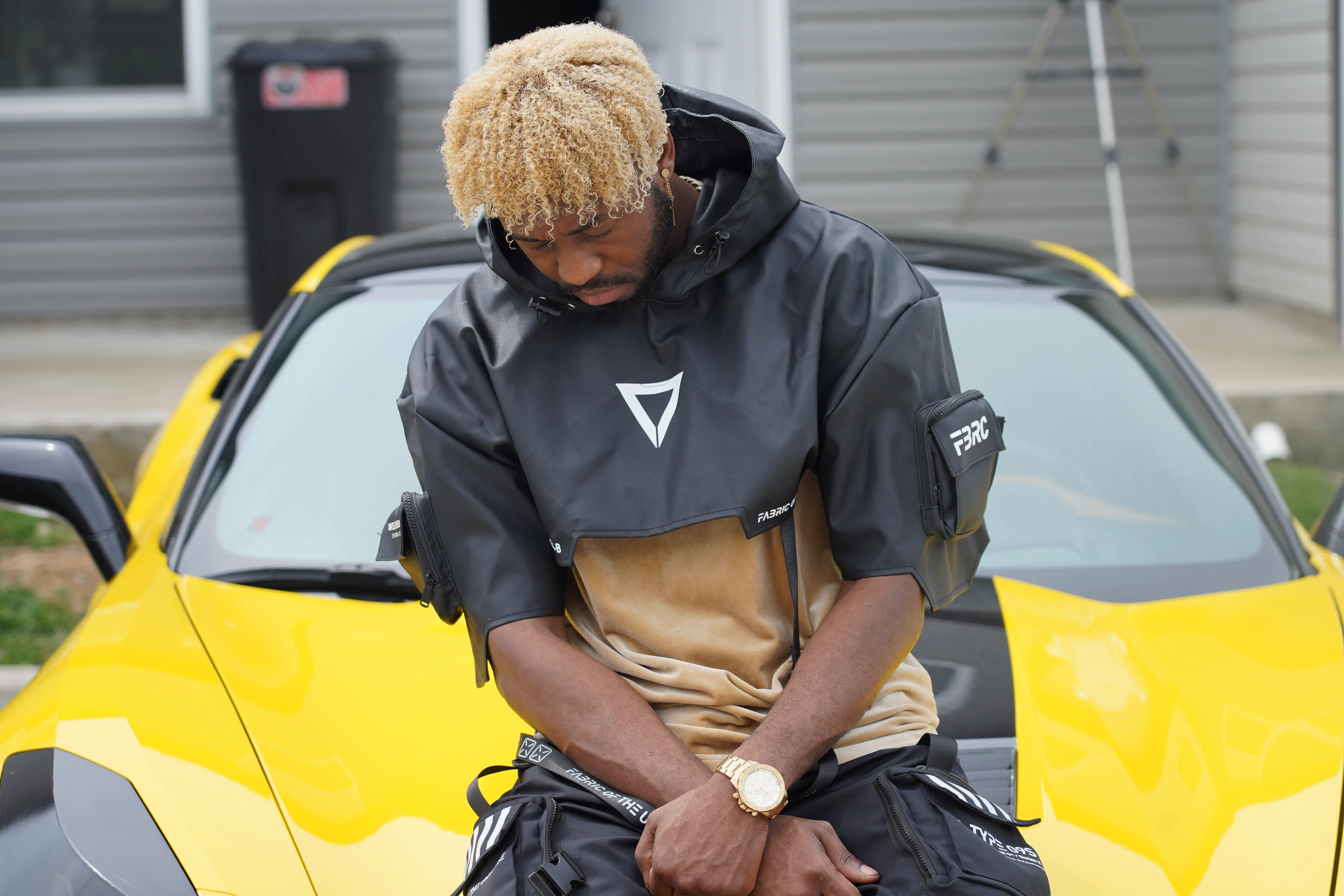 Alpha The Wolf seated on the yellow Corvette's hood with his head bowed and eyes downcast in a quiet, introspective pose, the grey tactical anorak and gold watch catching flat overcast light