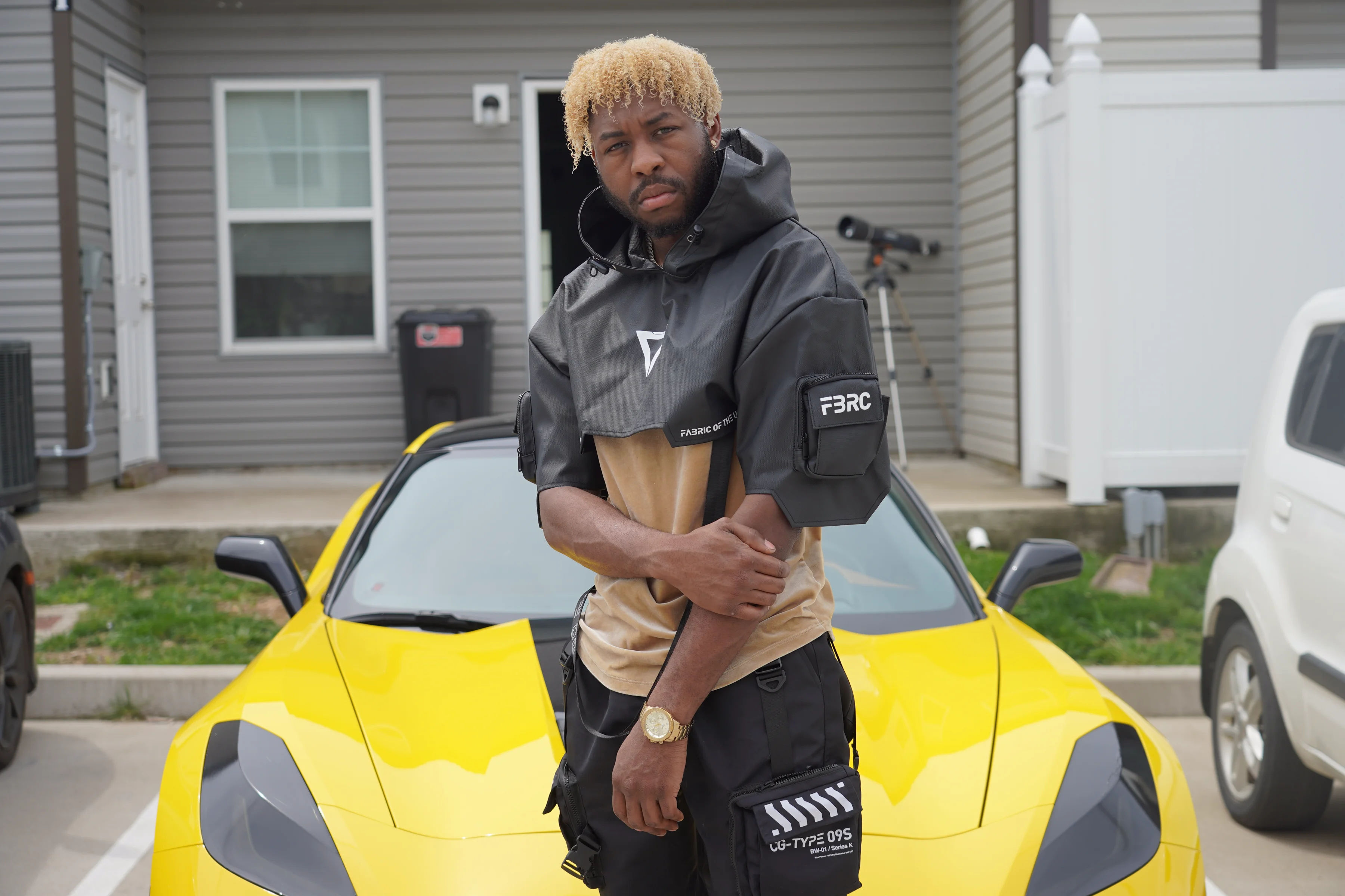 Alpha The Wolf standing in front of the yellow Corvette with arms crossed over his chest in a commanding stance, grey tactical anorak over a tan tee, gaze direct and confident