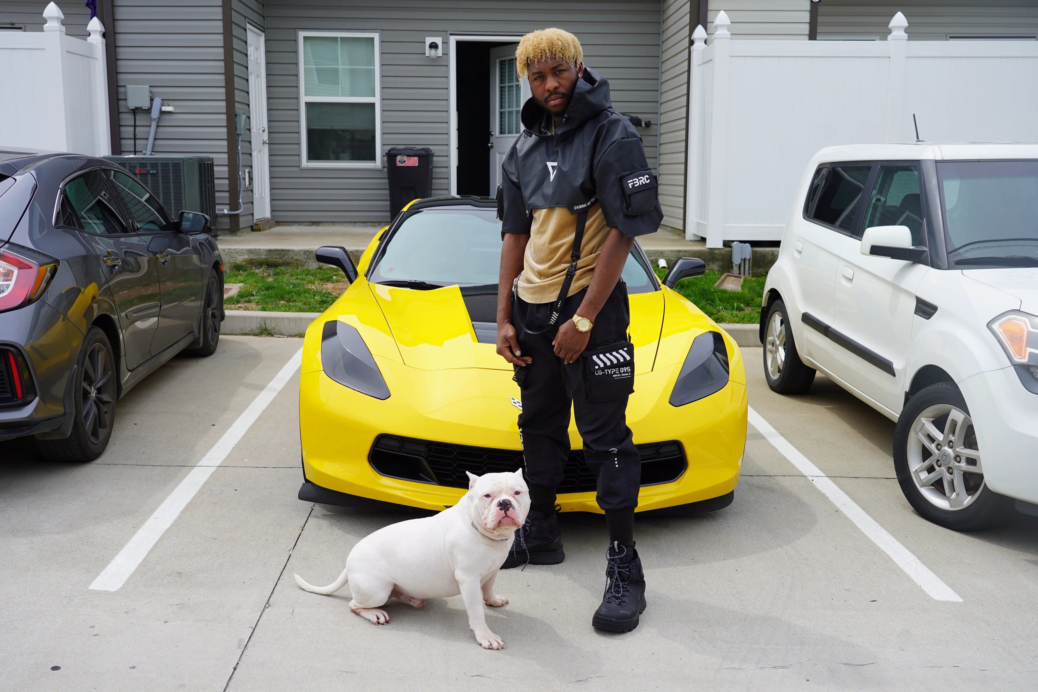 Alpha The Wolf standing upright in a parking lot in front of the yellow Corvette with the white American Bully sitting beside him, a full-length view of the grey tactical anorak, black cargo pants and chunky boots