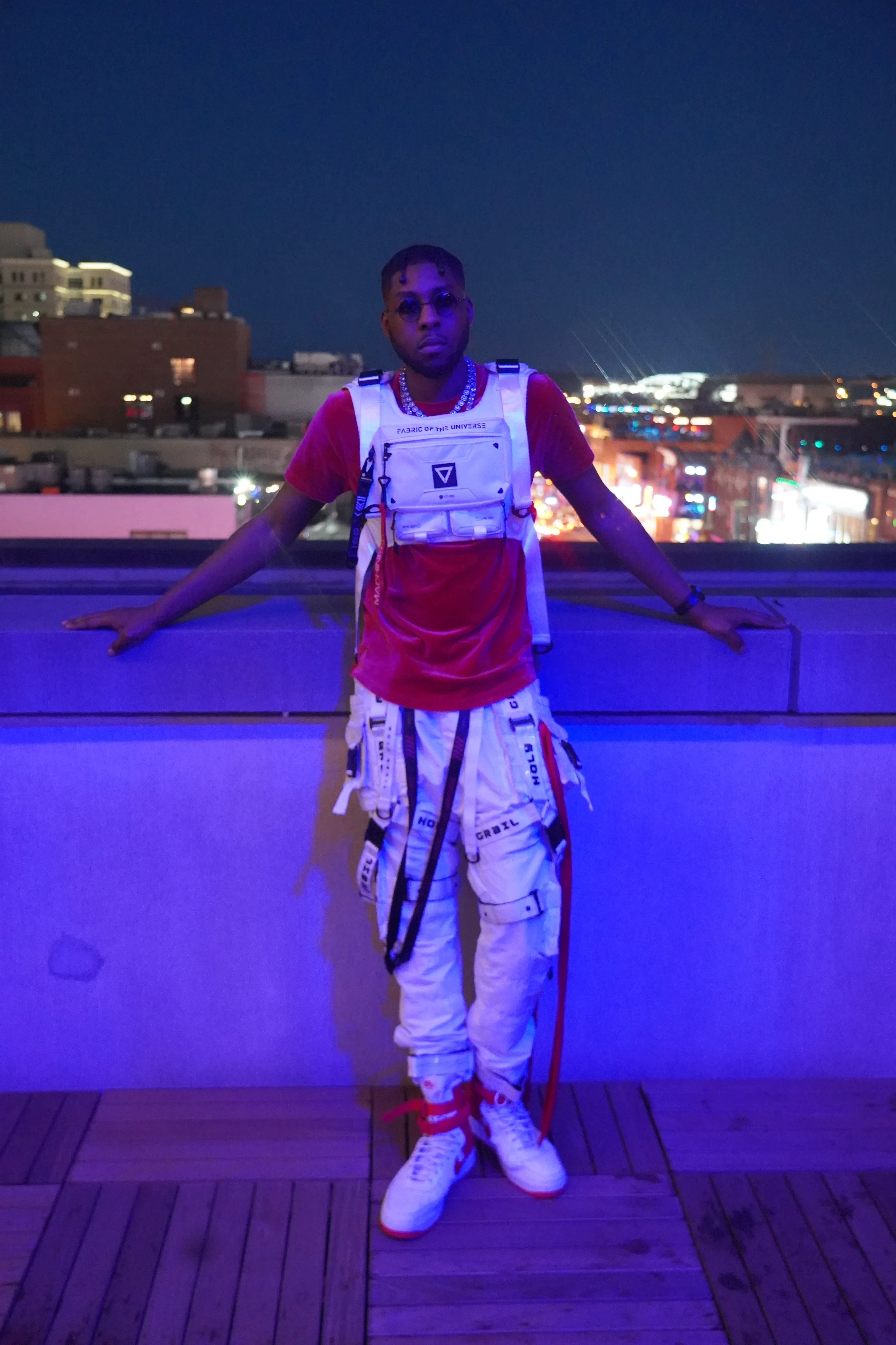 Alpha The Wolf standing full-length on a rooftop deck with both arms spread along a low parapet lit deep blue, wearing a red velour tee under a white tactical chest rig, white strapped cargo pants and red and white high-tops, the lit neon strip and city skyline behind him