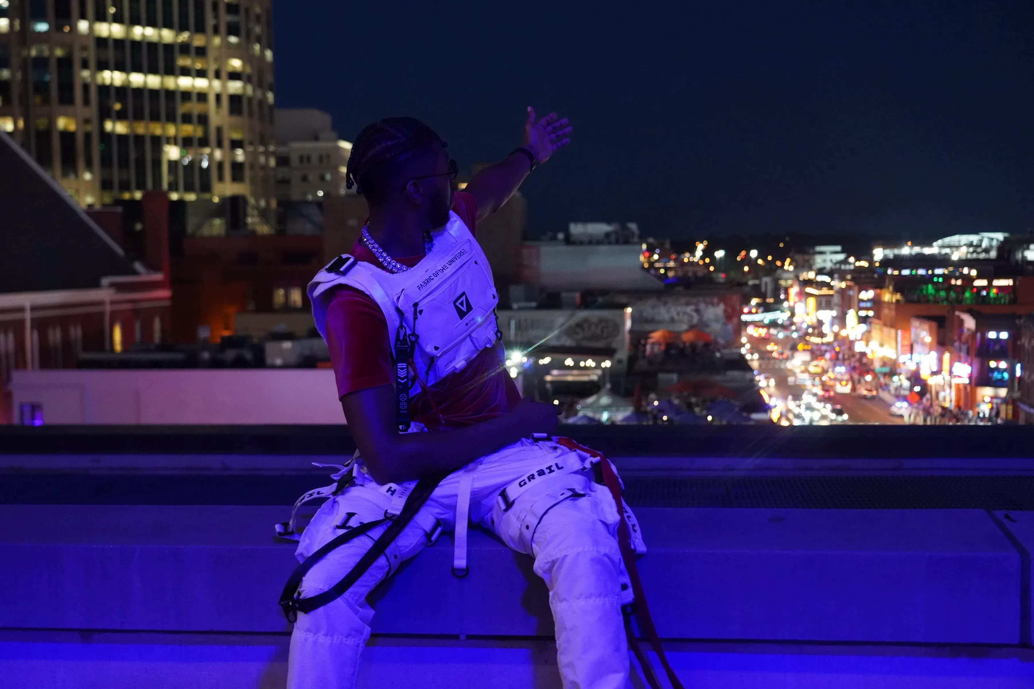 Alpha The Wolf sitting on the rooftop ledge with one arm thrown wide out to the side, head tilted, the whole lit street and its marquees spread out under him