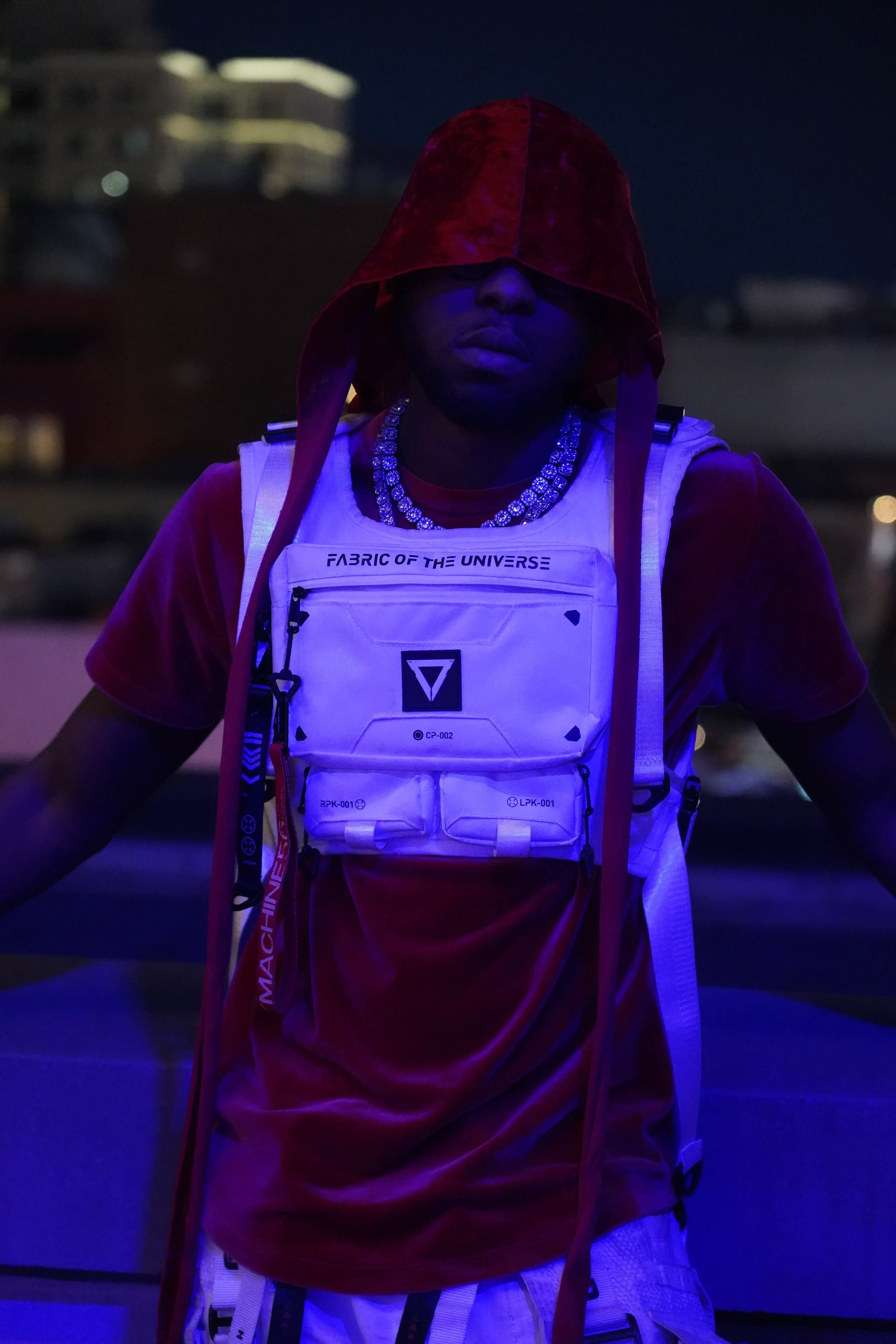 Alpha The Wolf in a close vertical frame with the red hood up shadowing his eyes, the white chest rig lit bright and lettered across the front, arms held out from his sides