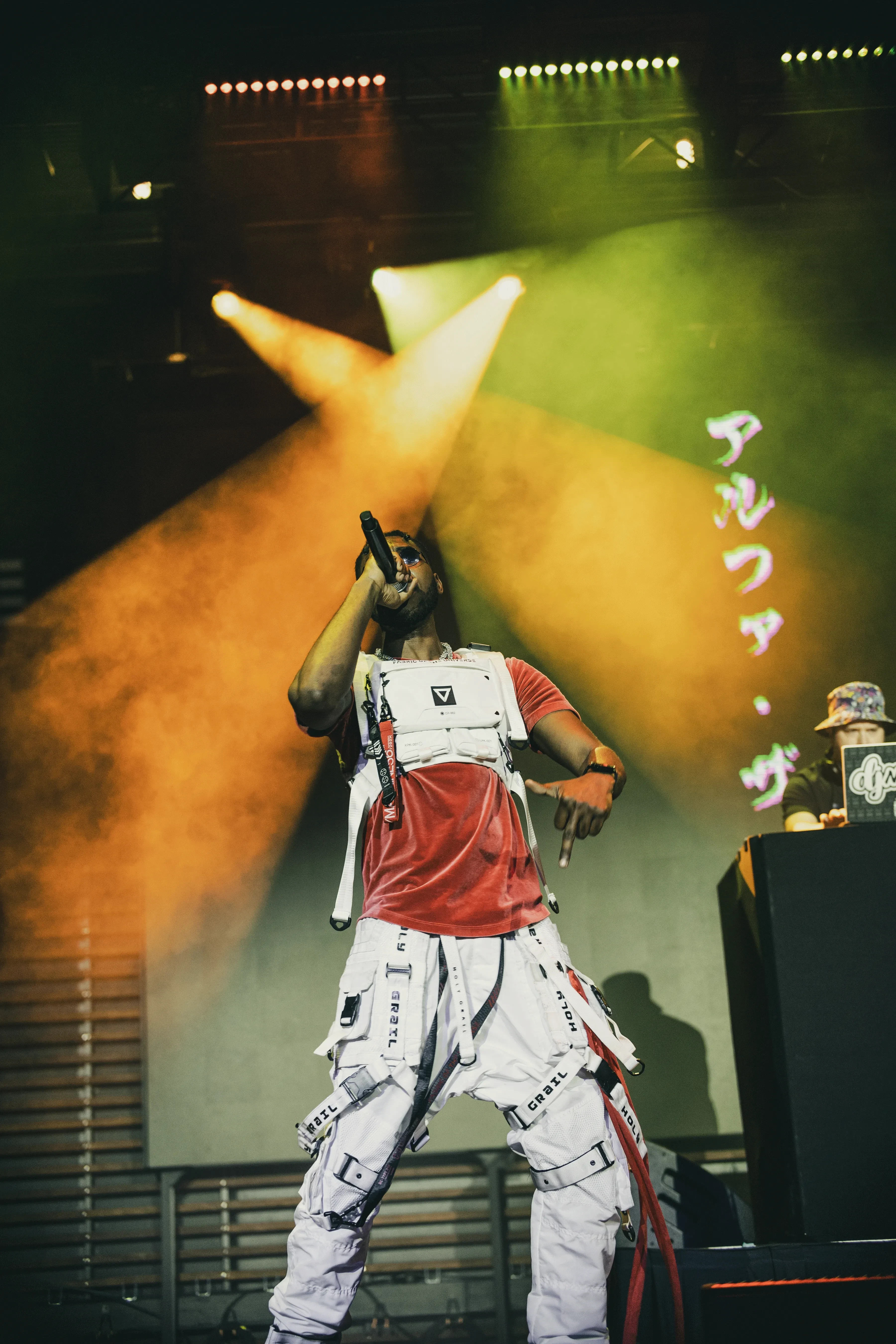 Alpha The Wolf shot from a low angle on stage with the microphone raised to his mouth and one arm lifted, orange spotlights cutting through haze and katakana characters projected on the backdrop
