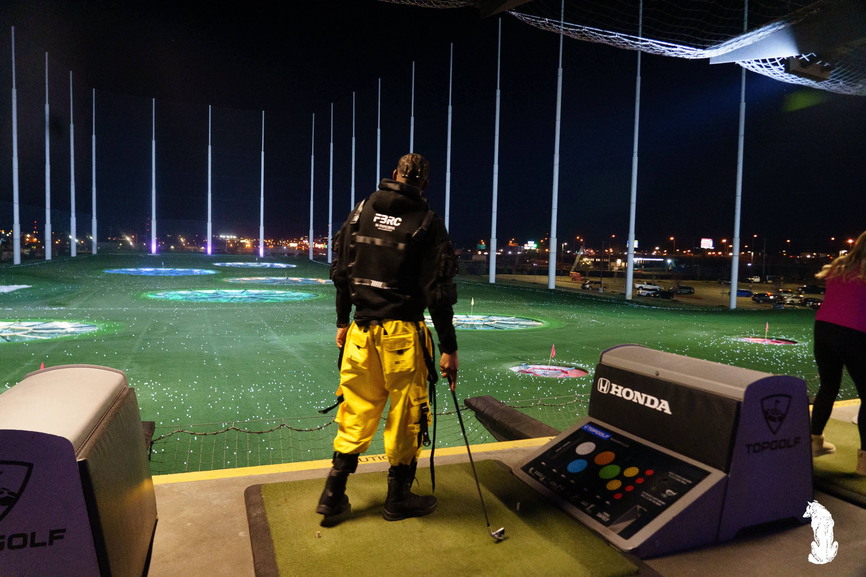 Alpha The Wolf at a Topgolf bay at night, back to the camera, holding a golf club at his side as he surveys the illuminated driving range, in a black tactical vest over a yellow hoodie and bright yellow cargo pants.