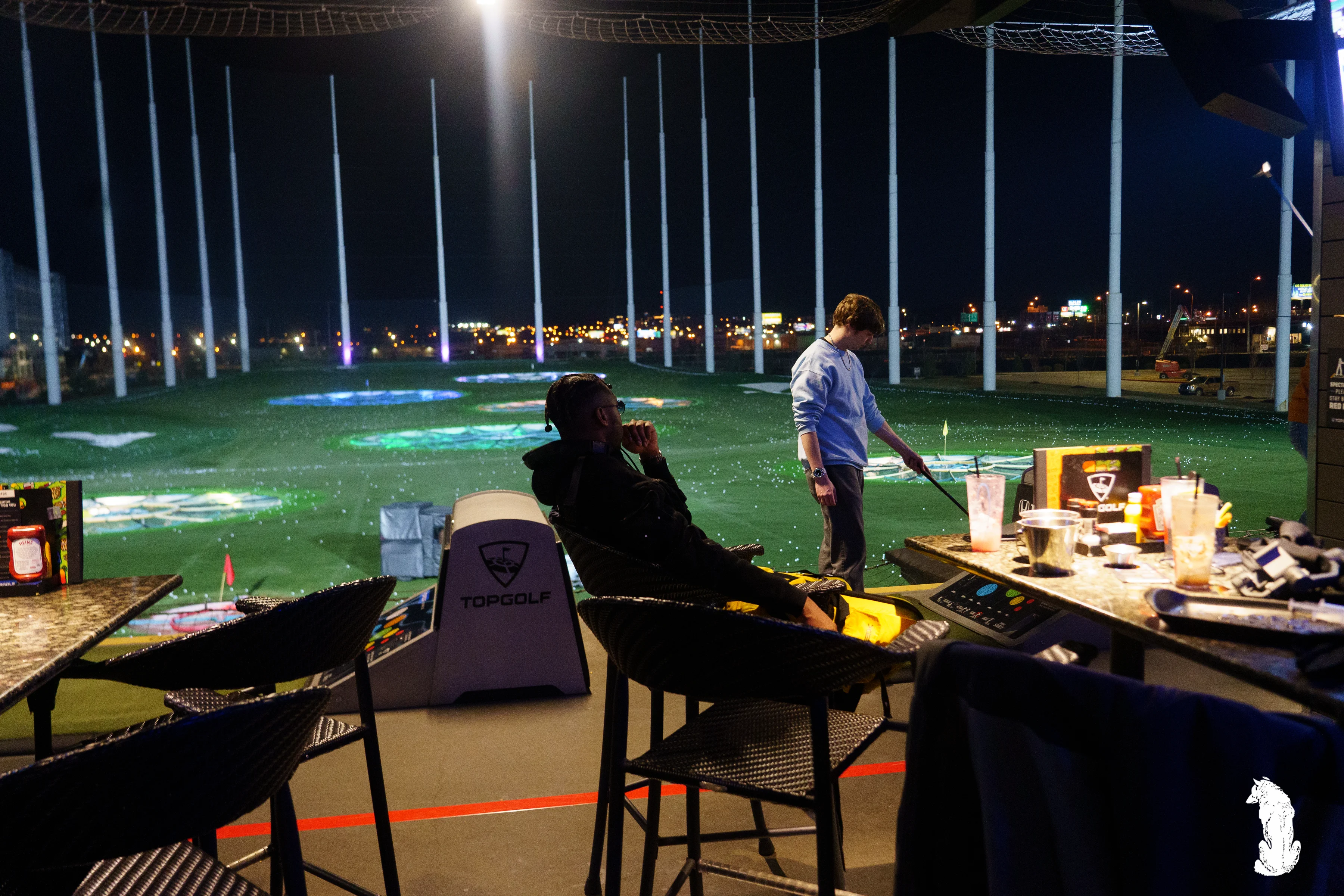 Alpha The Wolf reclined in a chair at the Topgolf bay, legs propped on the ledge, while a companion takes a swing at the tee and the glittering nighttime range spreads out behind.