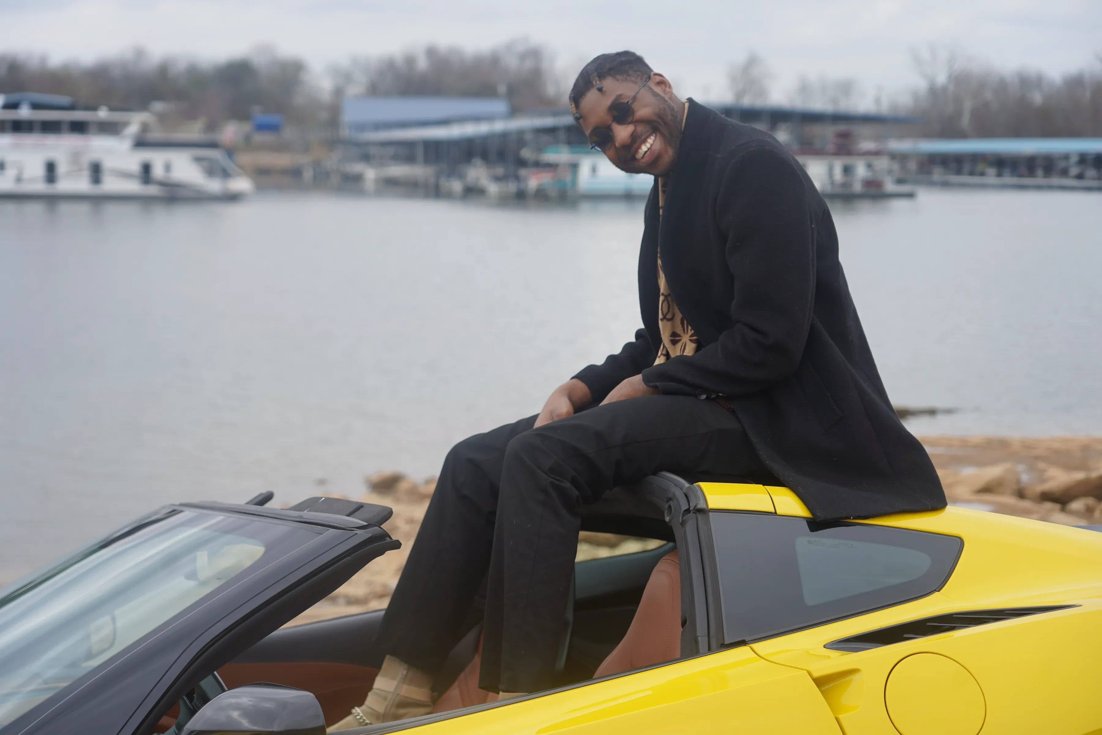 Alpha The Wolf smiling toward the camera from the roof of the yellow Corvette with one boot up on the door sill, the marina and a white houseboat on the water behind him.