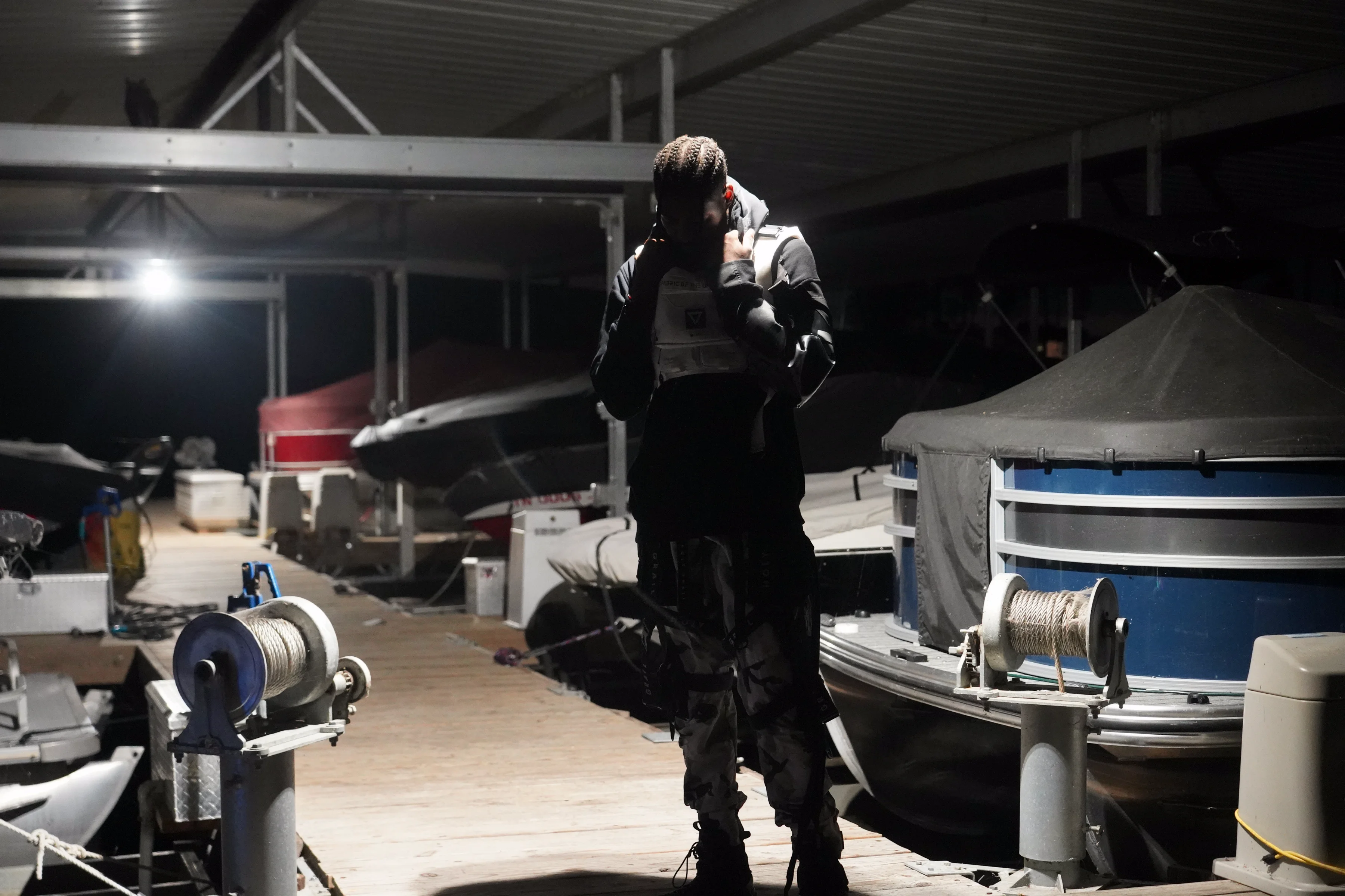 Alpha The Wolf standing alone on the dock between two covered pontoon boats, head bowed into his collar, the single overhead light behind him casting his all-black tactical outfit in silhouette