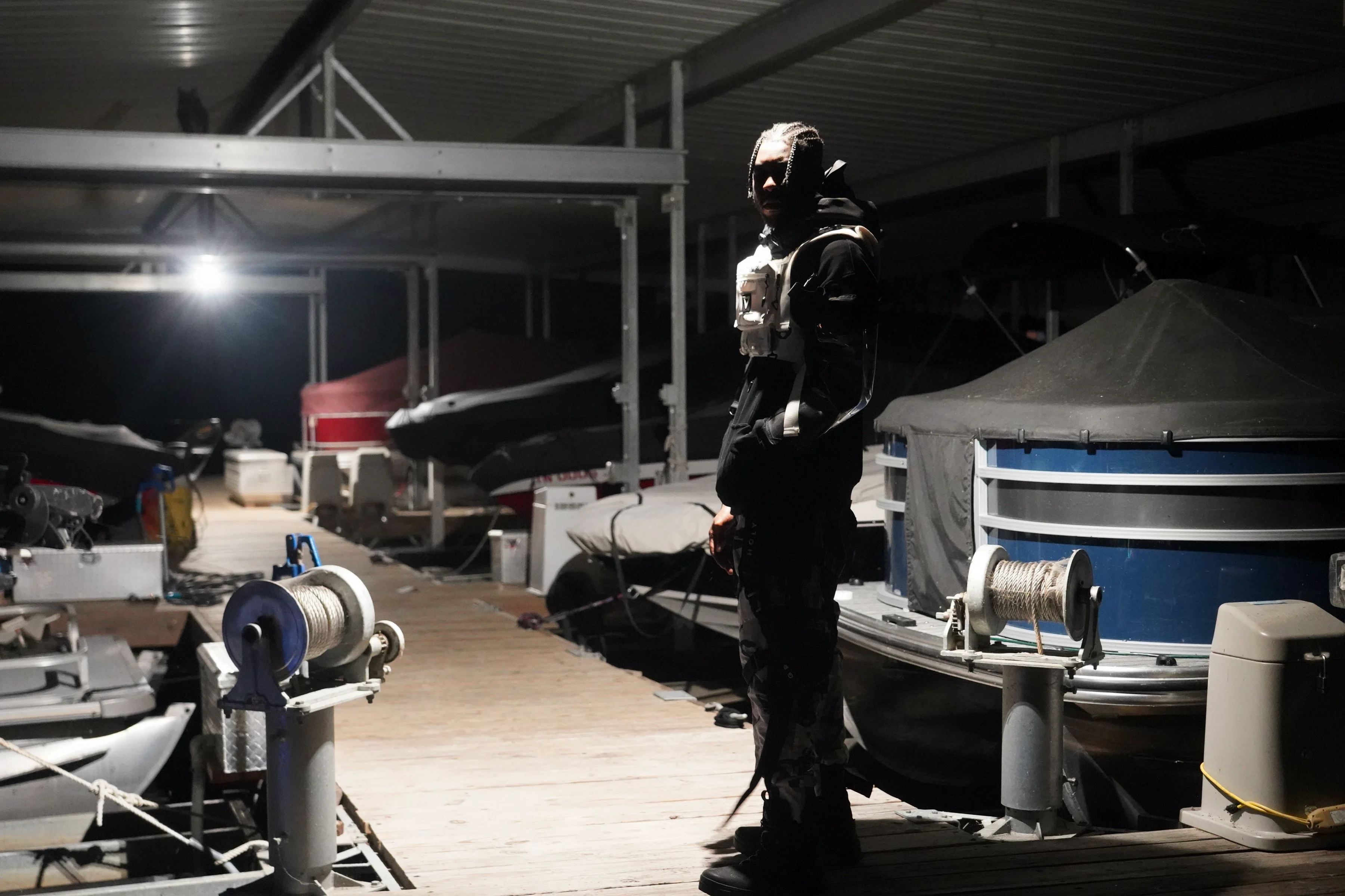 Alpha The Wolf standing tall on the marina dock in a full black tactical vest and cargo pants, glancing back over his shoulder toward the camera with quiet intensity, boats and dock equipment flanking him on both sides under dim warehouse lighting