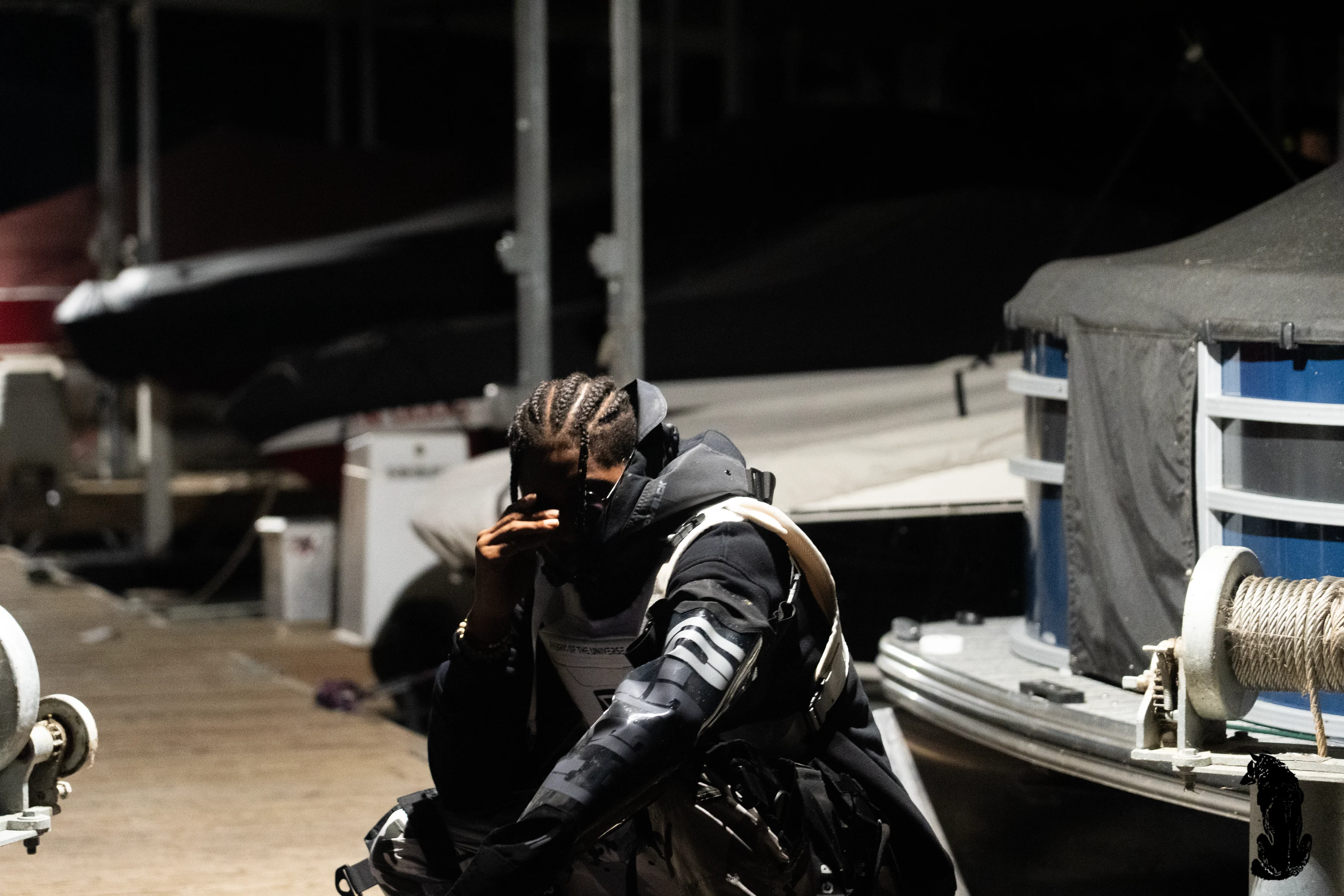 Alpha The Wolf in a close, low crouch on the dock, braided hair falling forward as he raises one hand to his face in a contemplative gesture, the black tactical vest visible in the tight frame