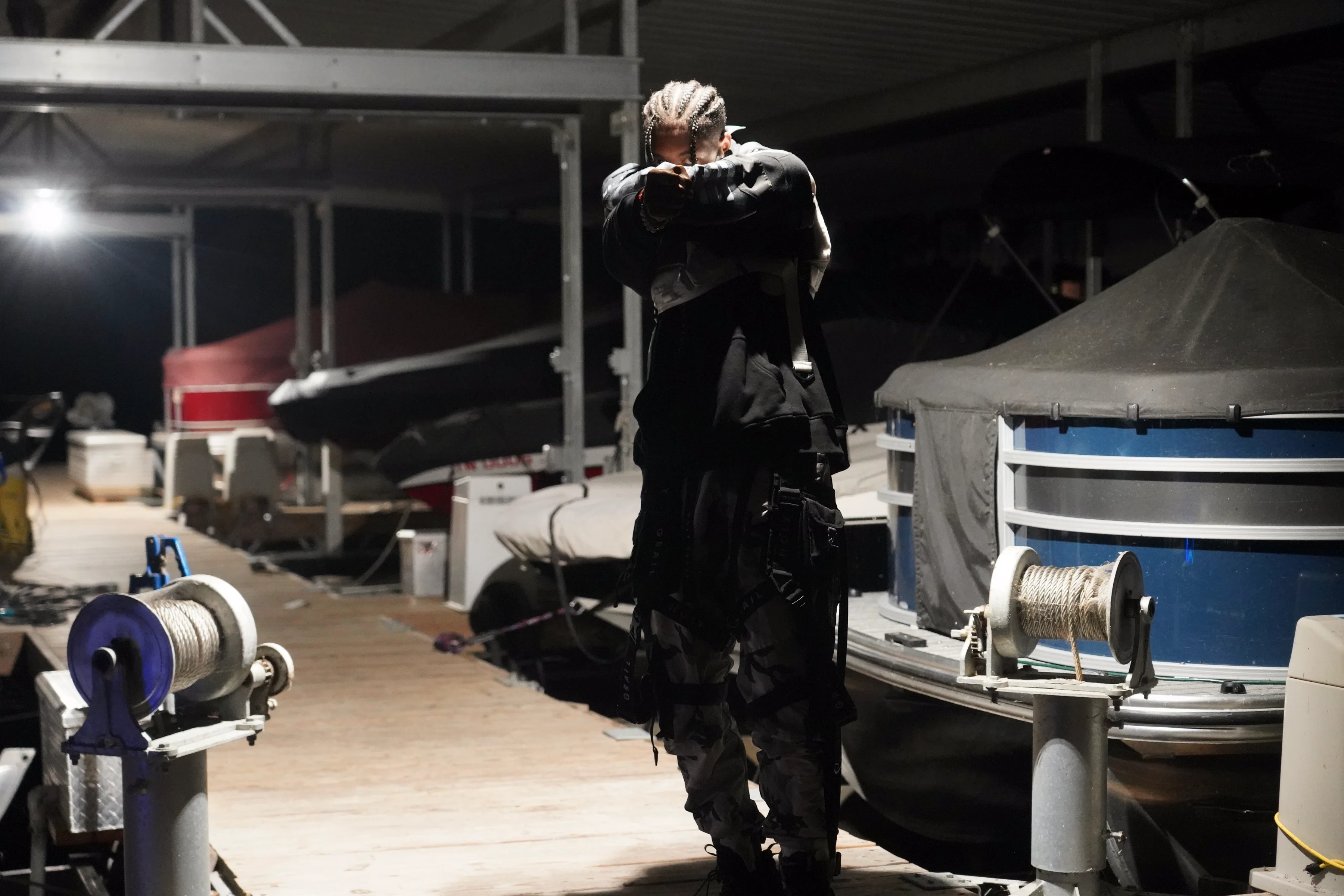 Alpha The Wolf shot from behind, back turned to the camera with his hand raised to the back of his head, the sprawling covered marina stretching before him under a solitary light