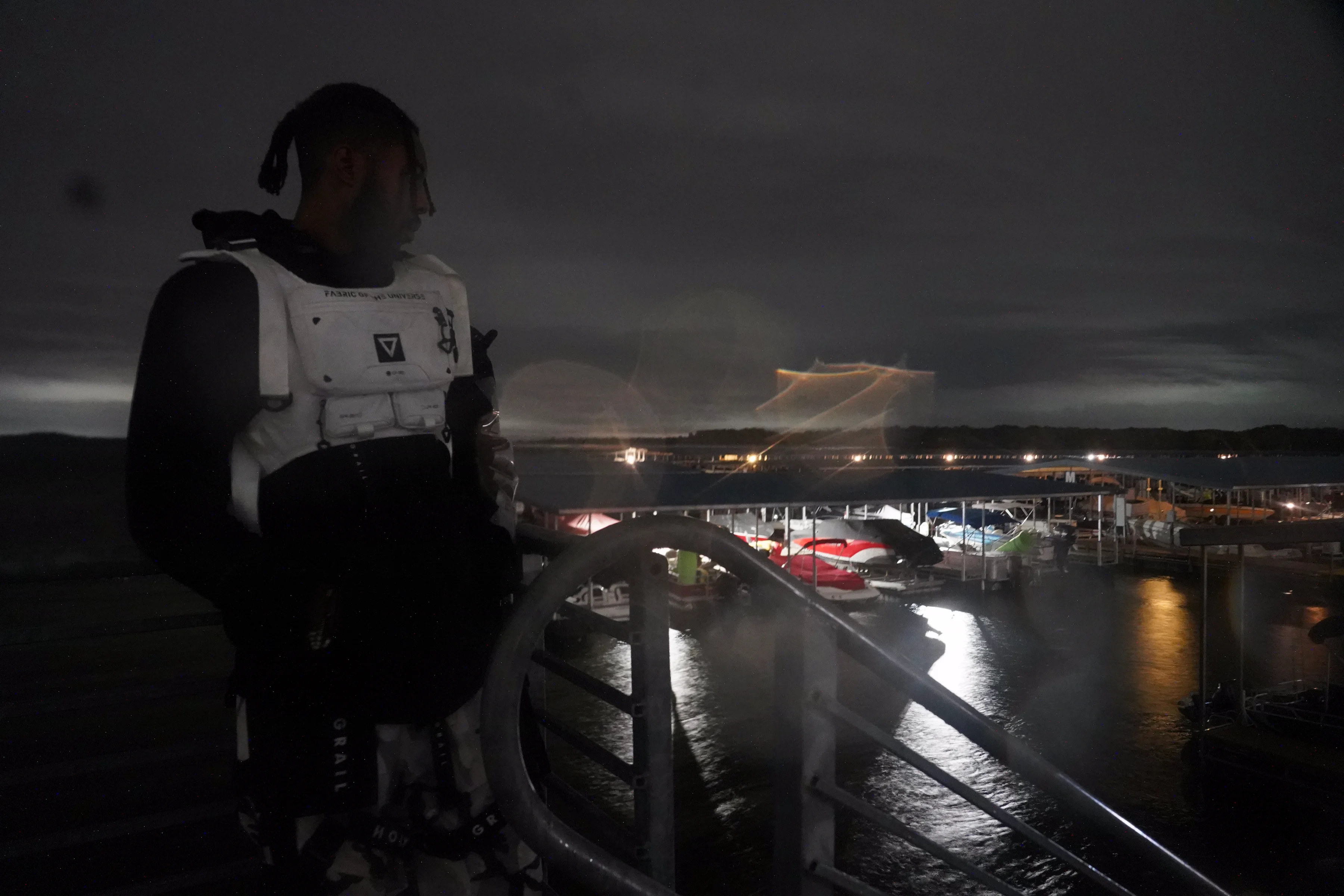 Alpha The Wolf standing at a rain-slicked upper deck railing overlooking a lit marina at night, profile to the camera in a white tactical vest, lightning illuminating the horizon above the treeline across the water in a dramatic wide shot