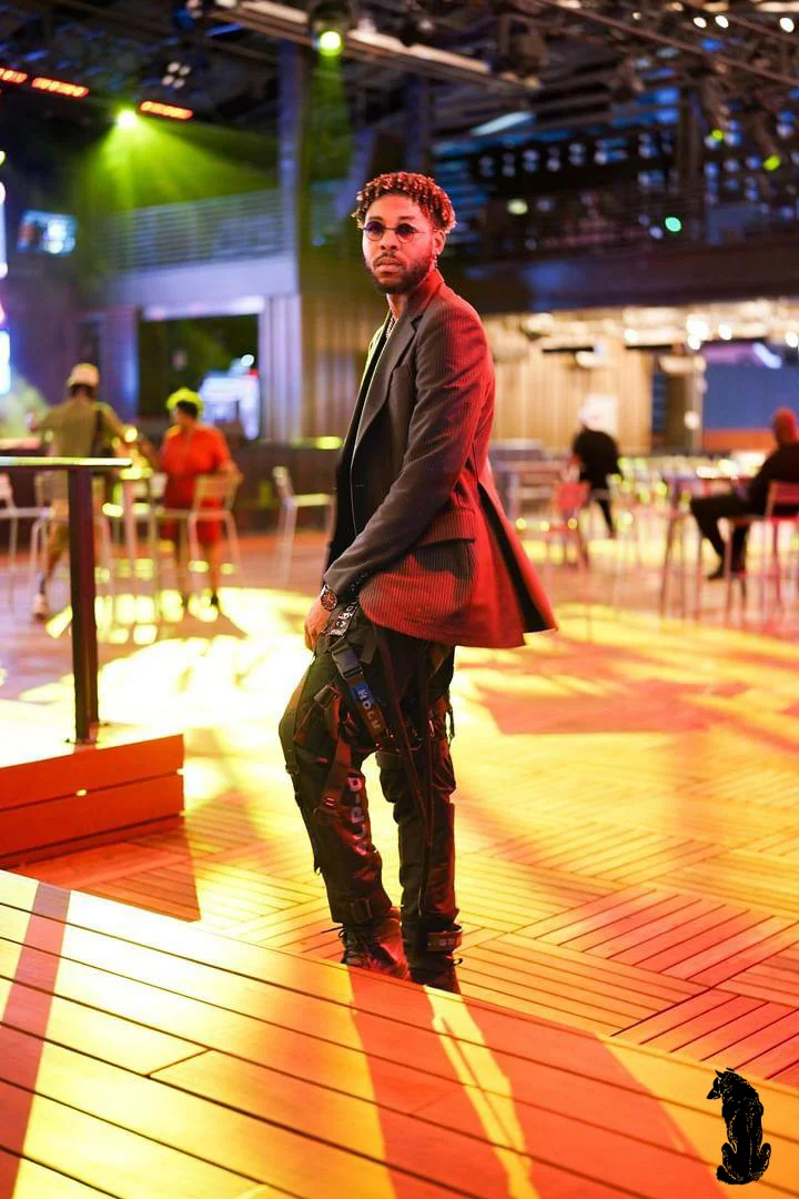 Alpha The Wolf standing full-length on a warmly lit wooden deck at night in a fitted charcoal blazer and tinted glasses, glancing back over his shoulder with cool detachment as venue patrons occupy the background