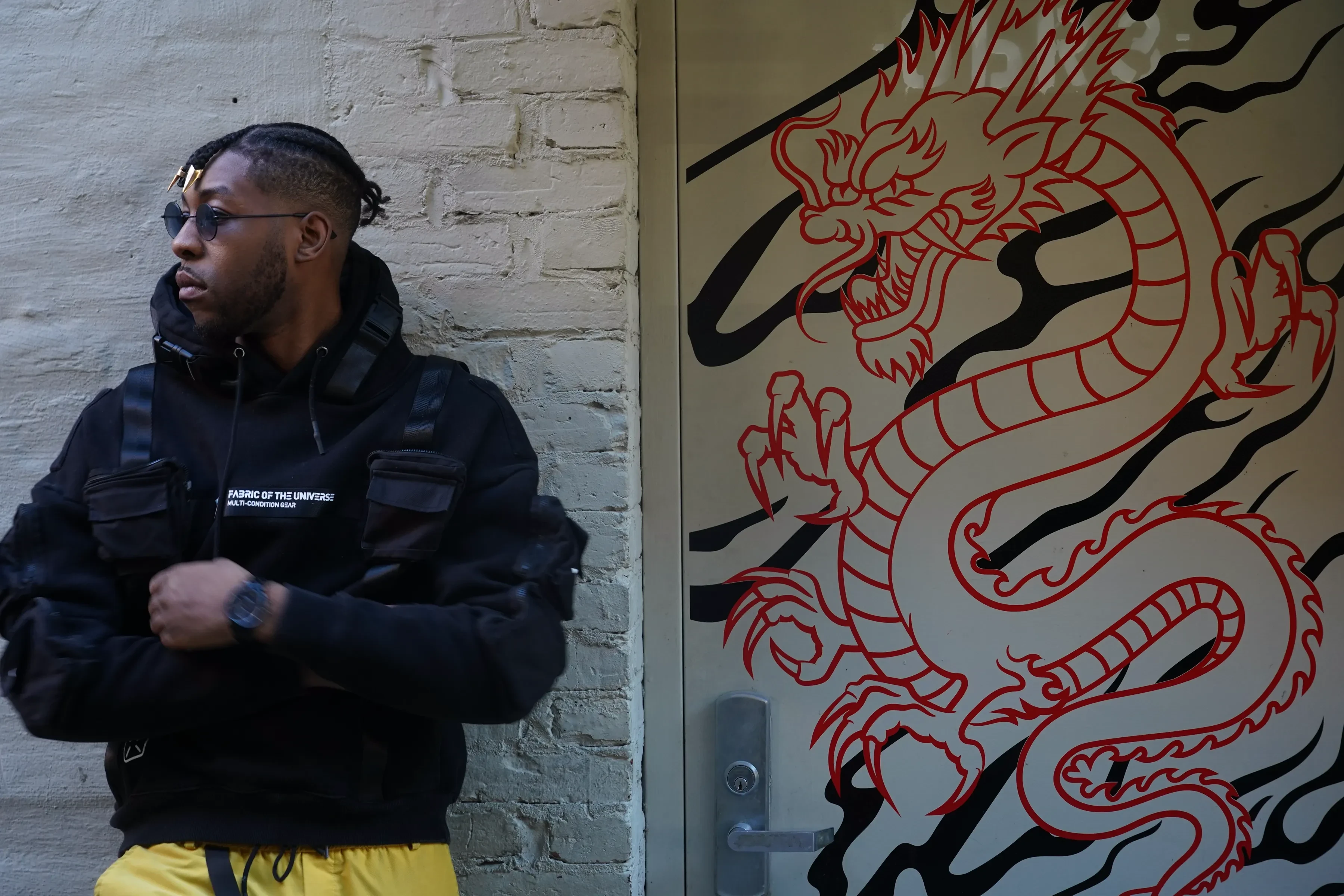 Alpha The Wolf in profile against the brick, one hand raised toward his chest, the black chest rigging and printed hoodie visible with the red dragon curling across the door to his right.