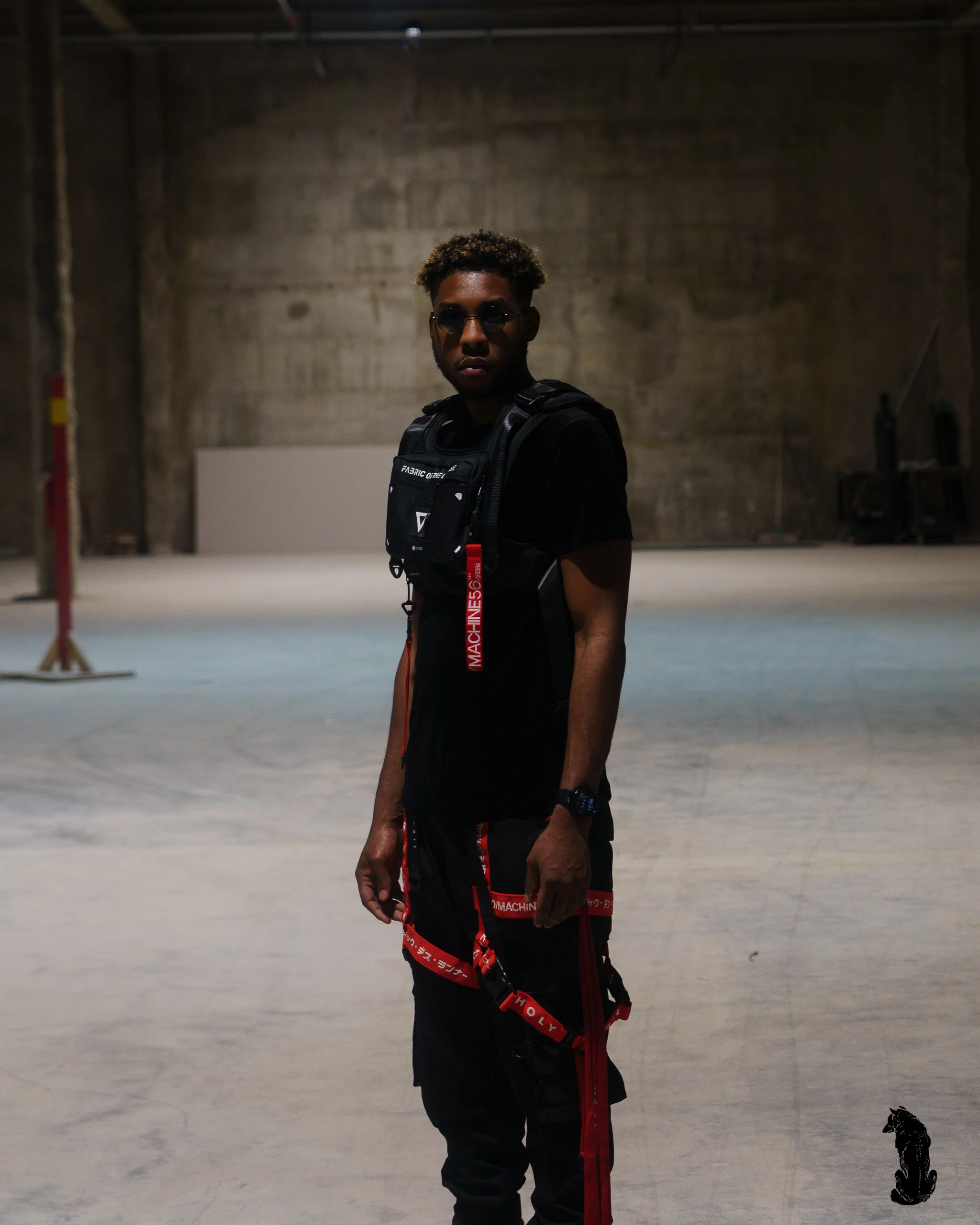 Alpha The Wolf standing alone in a vast empty concrete warehouse, staring into the camera in a black tactical chest rig over a black tee, red logo straps hanging from black cargo pants, blue-tinted frames on.