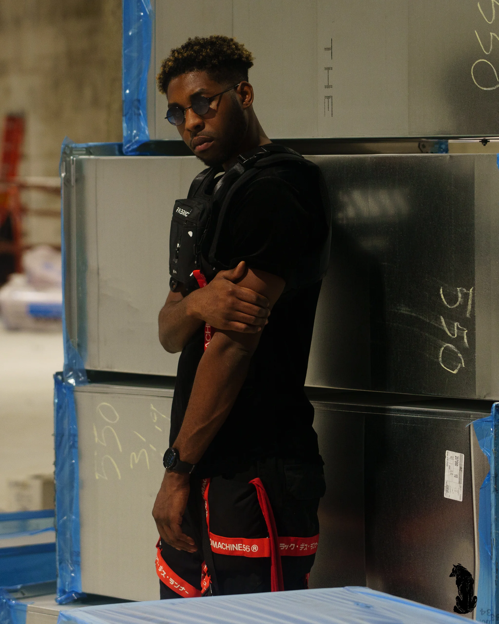 Alpha The Wolf leaning against a stack of metal panels chalked with numbers, arms crossed and gaze cutting sideways, in the black tactical rig and red-strapped cargo pants.
