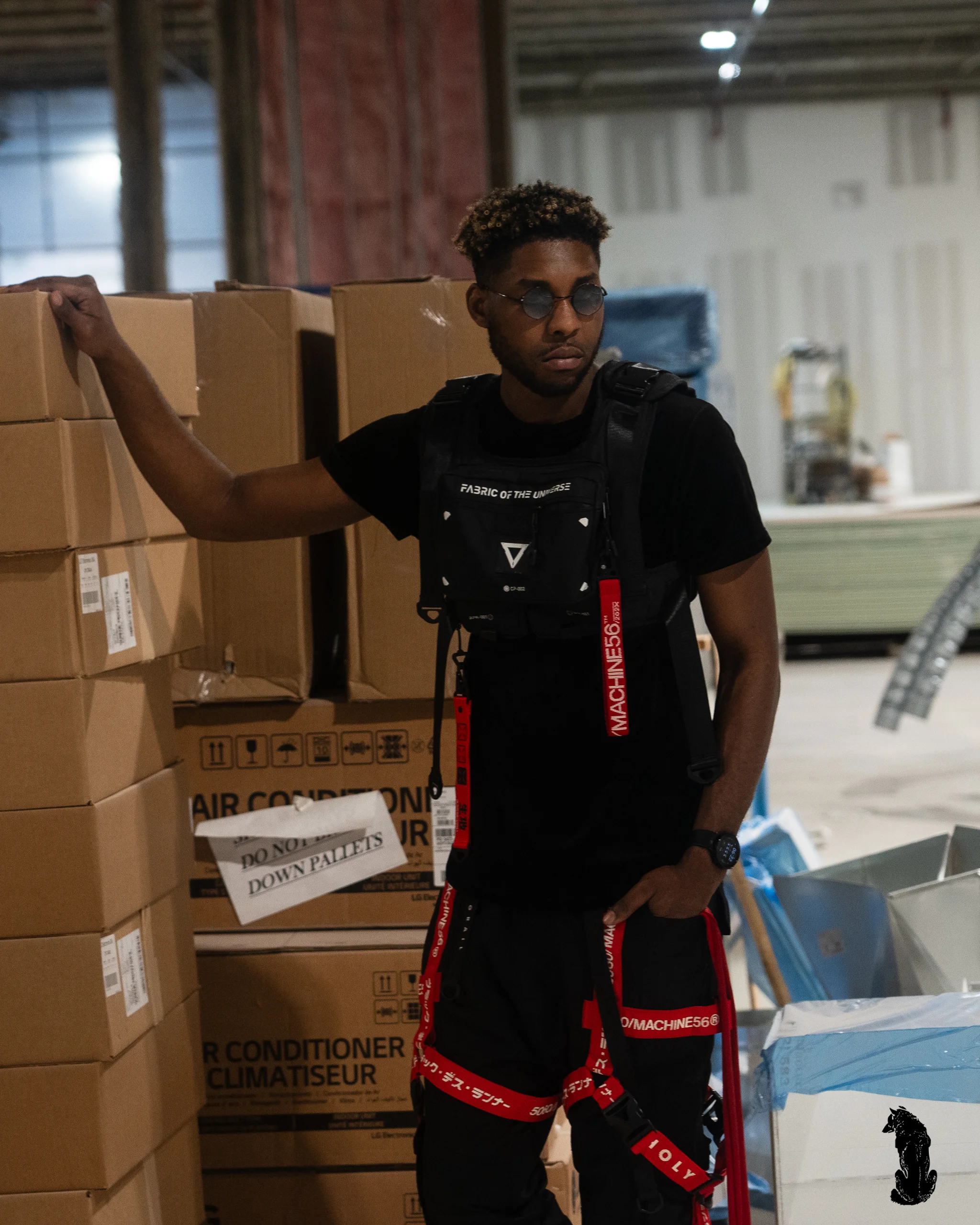 Alpha The Wolf posed beside a tall column of stacked cardboard boxes in the warehouse, one hand resting on the top box, red cargo straps and chest rig prominent in the warm ambient light.