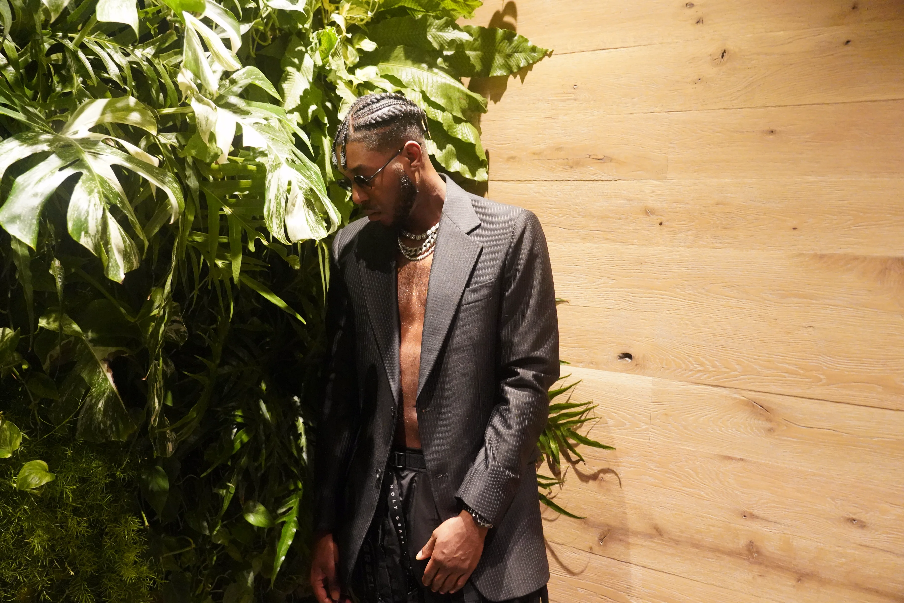 Alpha The Wolf standing beside a lush living plant wall of monstera and tropical foliage, head bowed in quiet introspection, an open charcoal blazer over a bare chest with layered chains and tactical cargo pants
