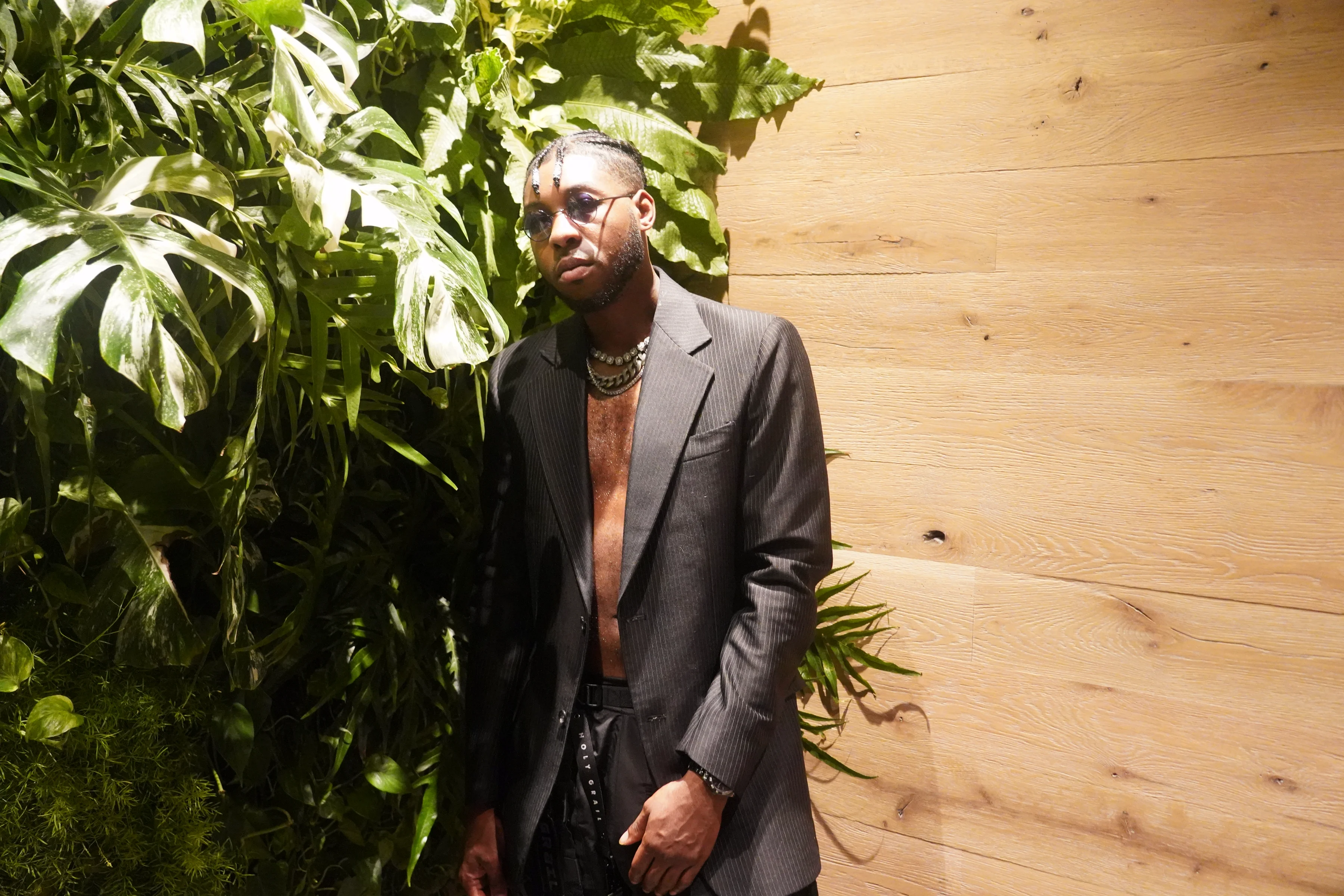 Alpha The Wolf leaning against the wood-panel wall next to the tropical plant installation, staring directly into camera with a cool, guarded expression, an open charcoal blazer and chain necklaces