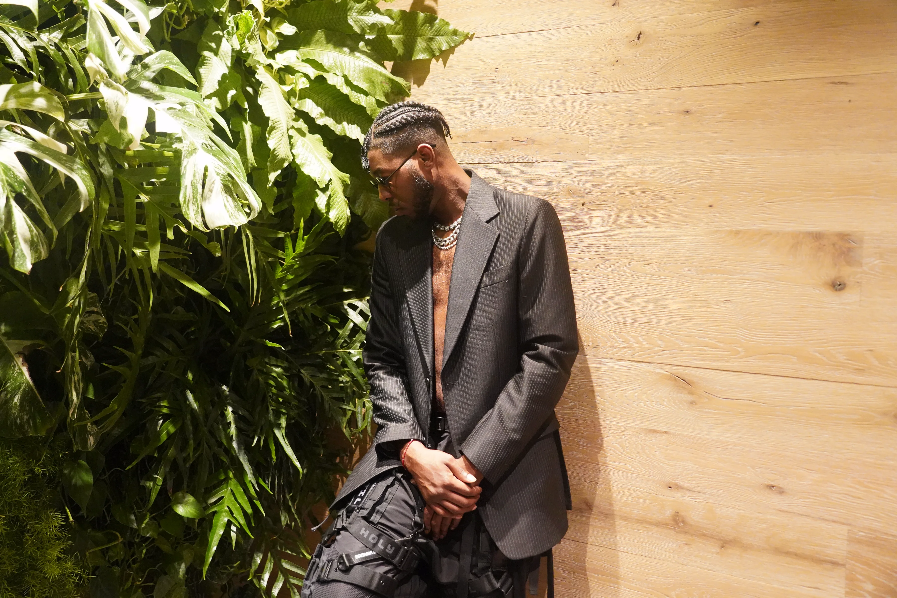 Alpha The Wolf bowing his head forward with hands clasped low, framed by dense monstera leaves pressing in from the left, an open charcoal blazer and tactical cargo pants