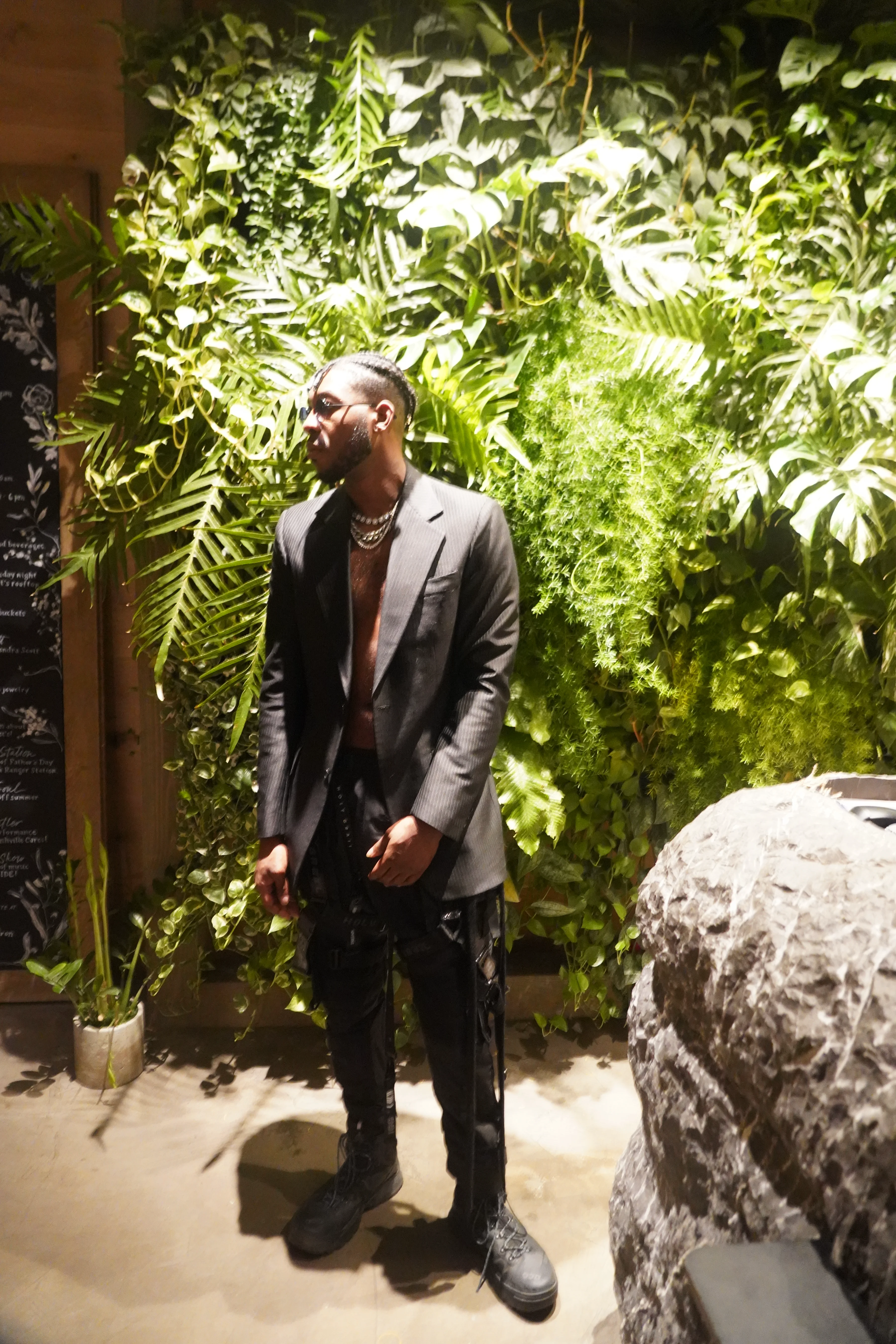 Alpha The Wolf with his face tilted up and eyes closed as if soaking in the atmosphere, green foliage lit from behind creating a luminous wall of leaves, open blazer and combat boots