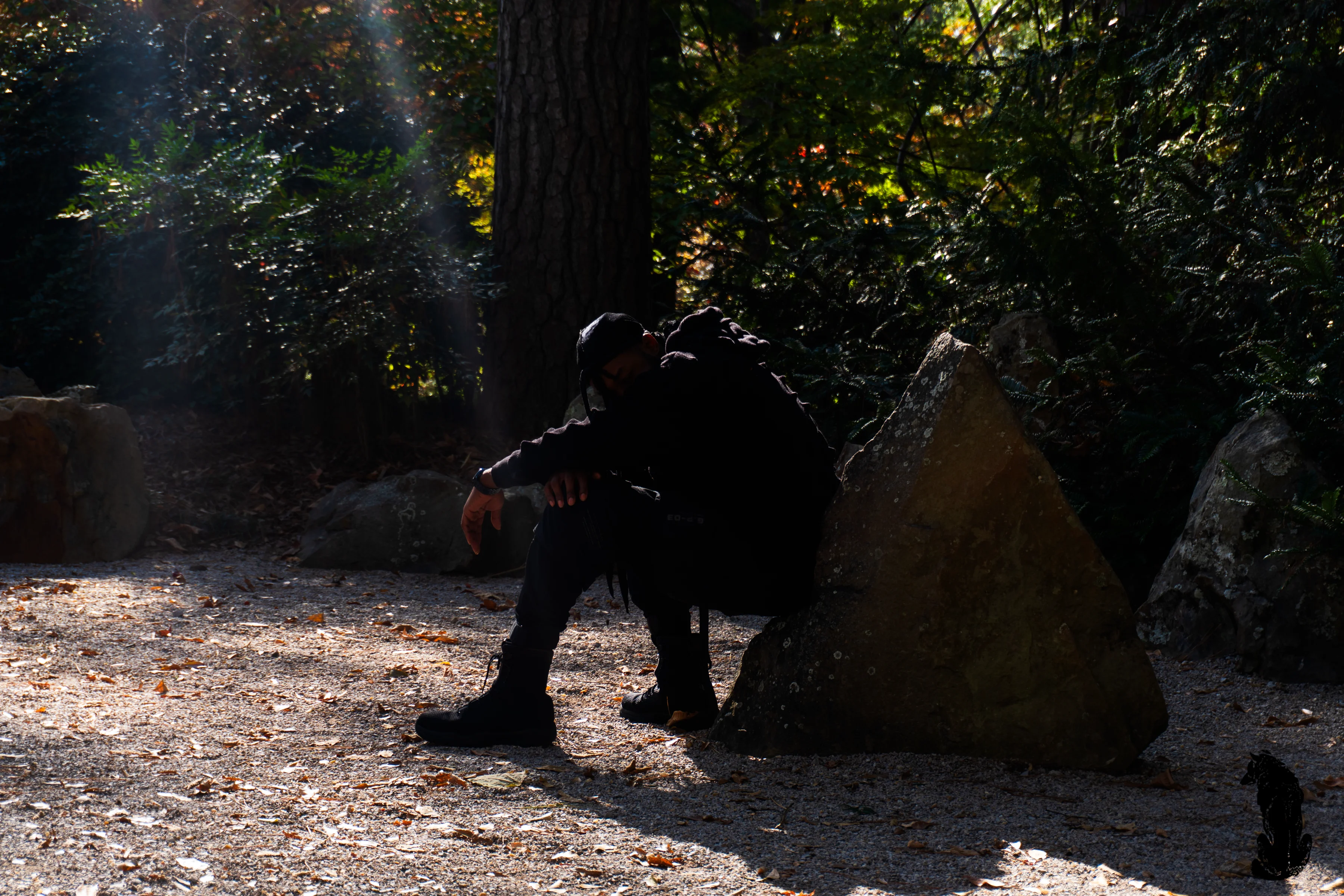 Alpha The Wolf standing on a stone pathway with his back to a clay tile-capped earthen wall, locs spilling down his back, arms crossed and head bowed, red maple trees in full autumn colour above the wall