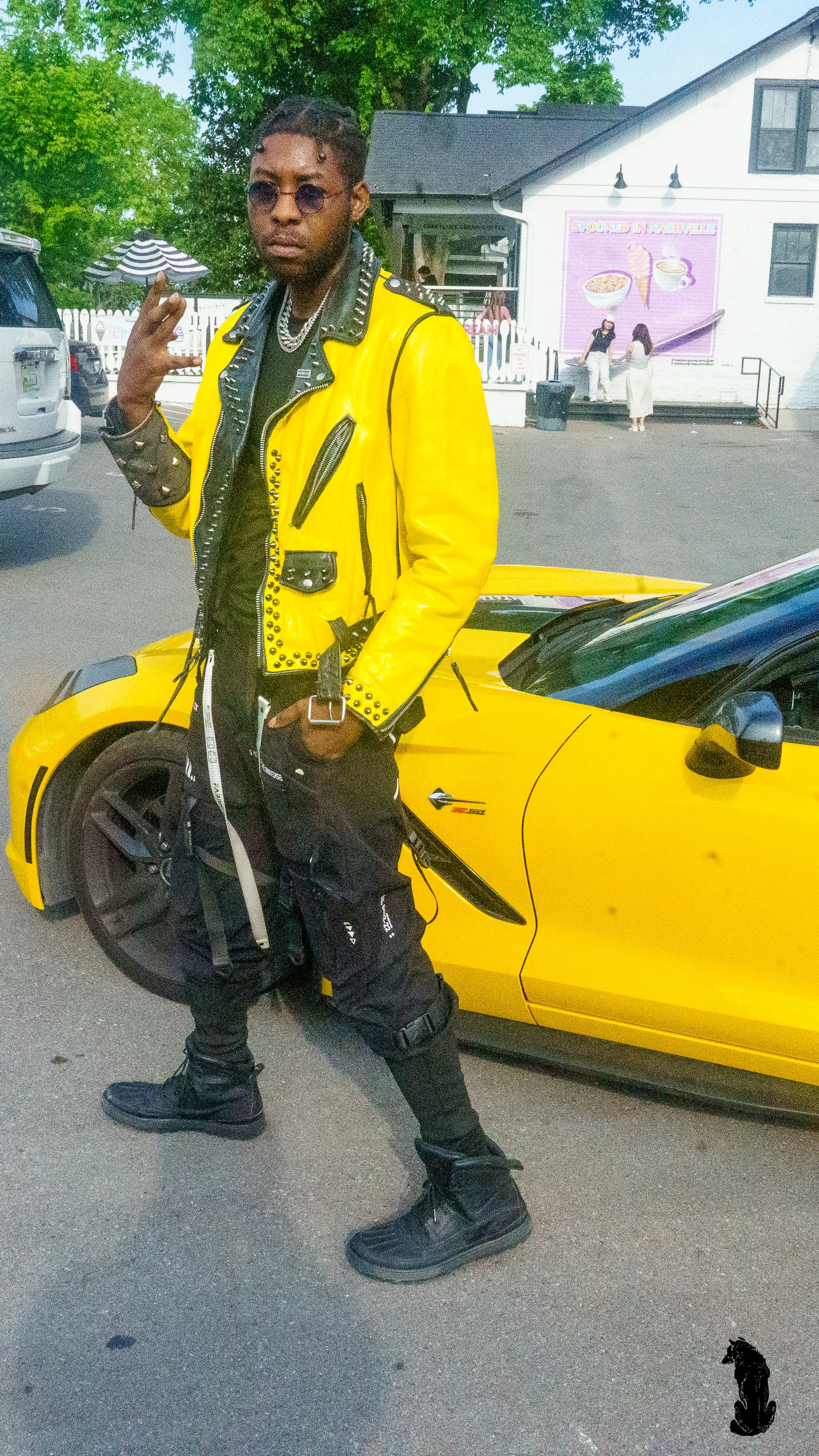 Alpha The Wolf standing in front of a matching yellow Corvette throwing a hand gesture toward the camera, a yellow studded leather moto jacket over a dark tee, black strapped cargo pants and boots, outside a white market building.