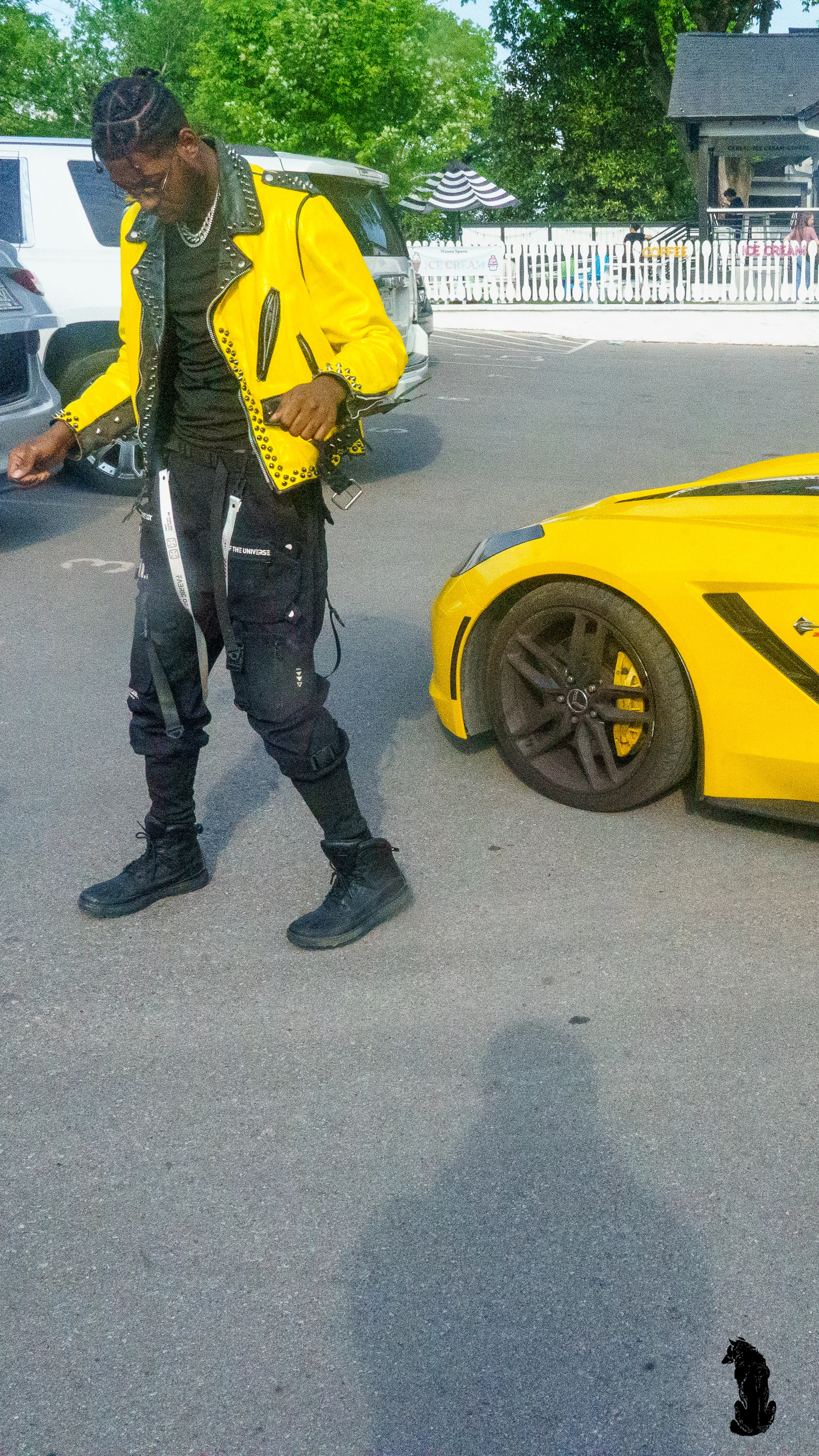 Alpha The Wolf walking in stride beside the yellow Corvette, head slightly bowed with a subtle smile, the car's low-slung body and chrome rims filling the right of the frame.