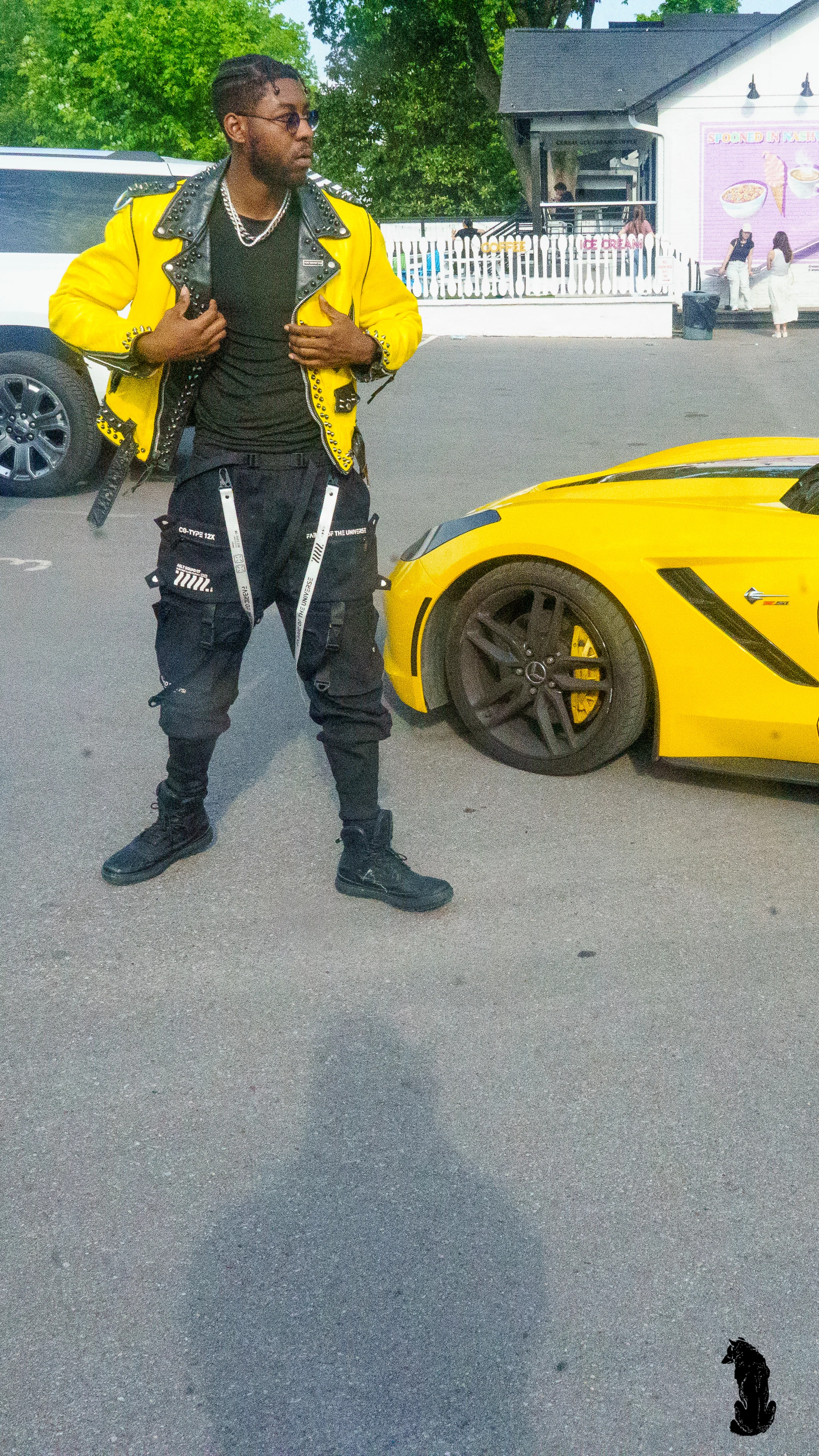 Alpha The Wolf standing beside the rear quarter of the yellow Corvette, gripping his lapel and gazing off-frame, shot from a low angle that emphasizes his height against a bright summer sky.