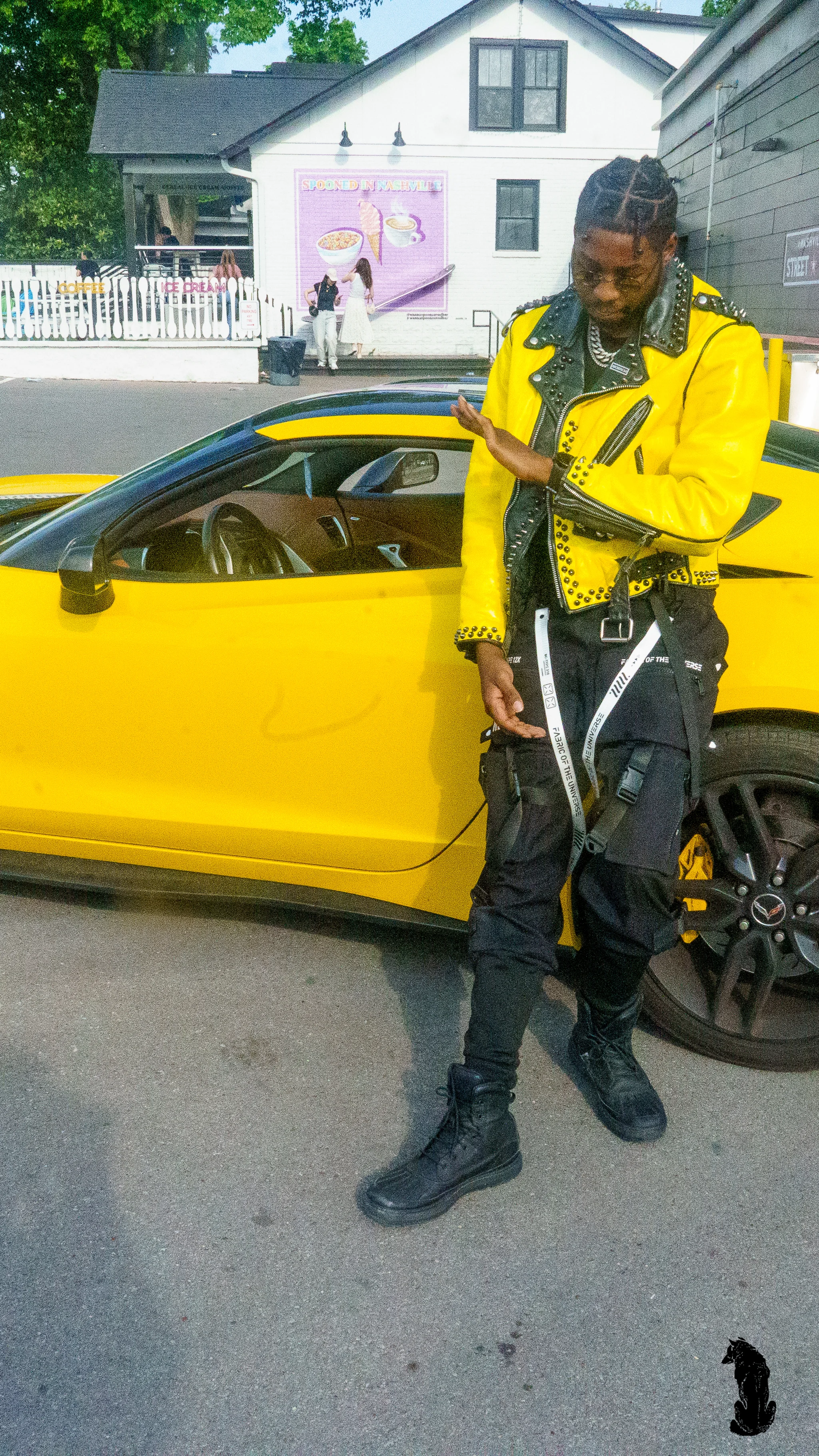 Alpha The Wolf leaning against the open door of the yellow Corvette, looking down with a relaxed smirk and adjusting his jacket cuff, a mural-covered white building in the soft background.