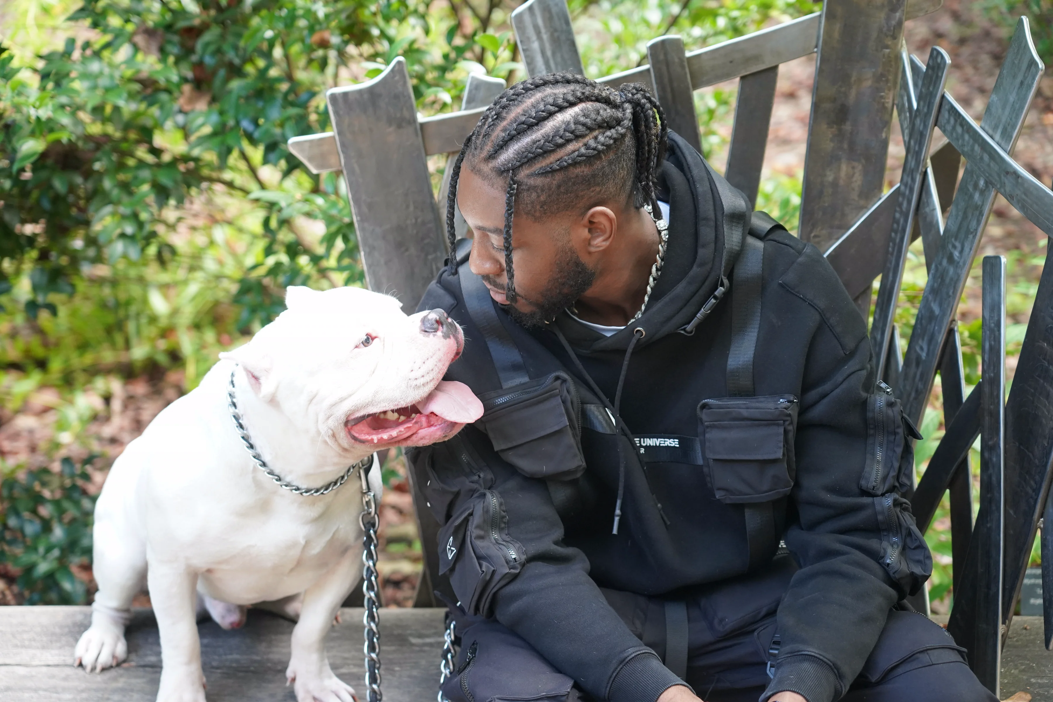Alpha The Wolf leaning in to nuzzle his white American Bully on a wooden bench in a garden setting, a tender candid moment with both faces nearly touching, soft light through the trees