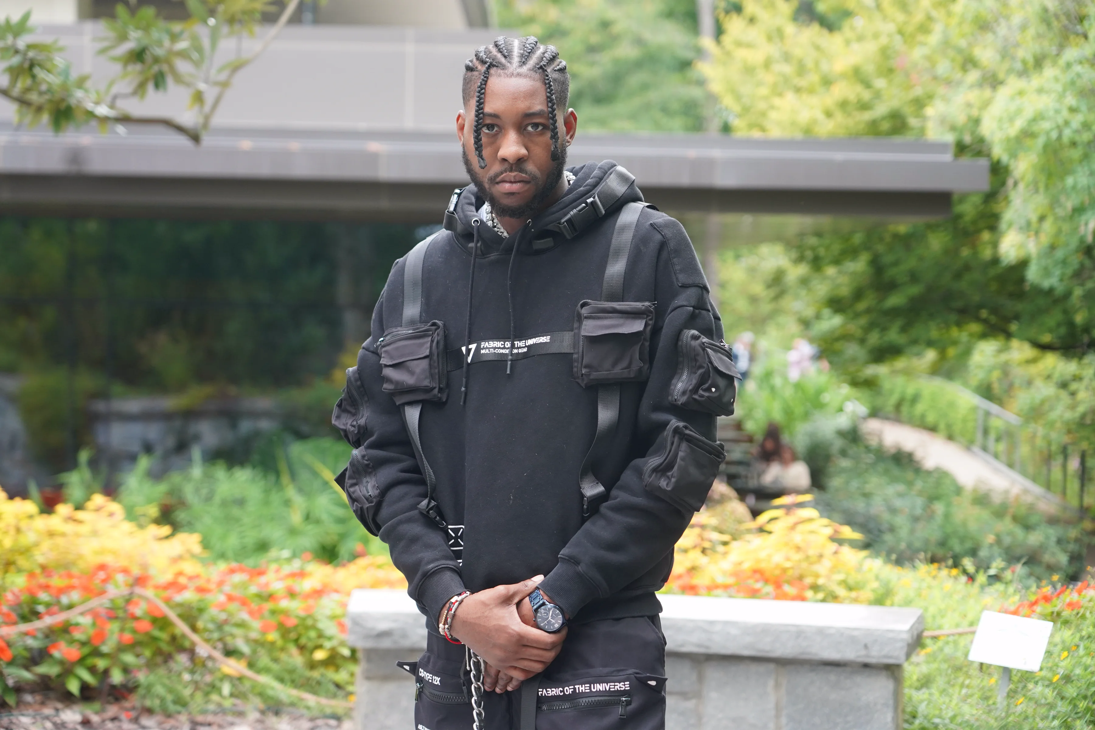 Alpha The Wolf in a direct-facing portrait against a garden backdrop, hands clasped in front, an all-black multi-pocket tactical hoodie and chest rig, colourful flower beds and a glass pavilion structure blurred behind him