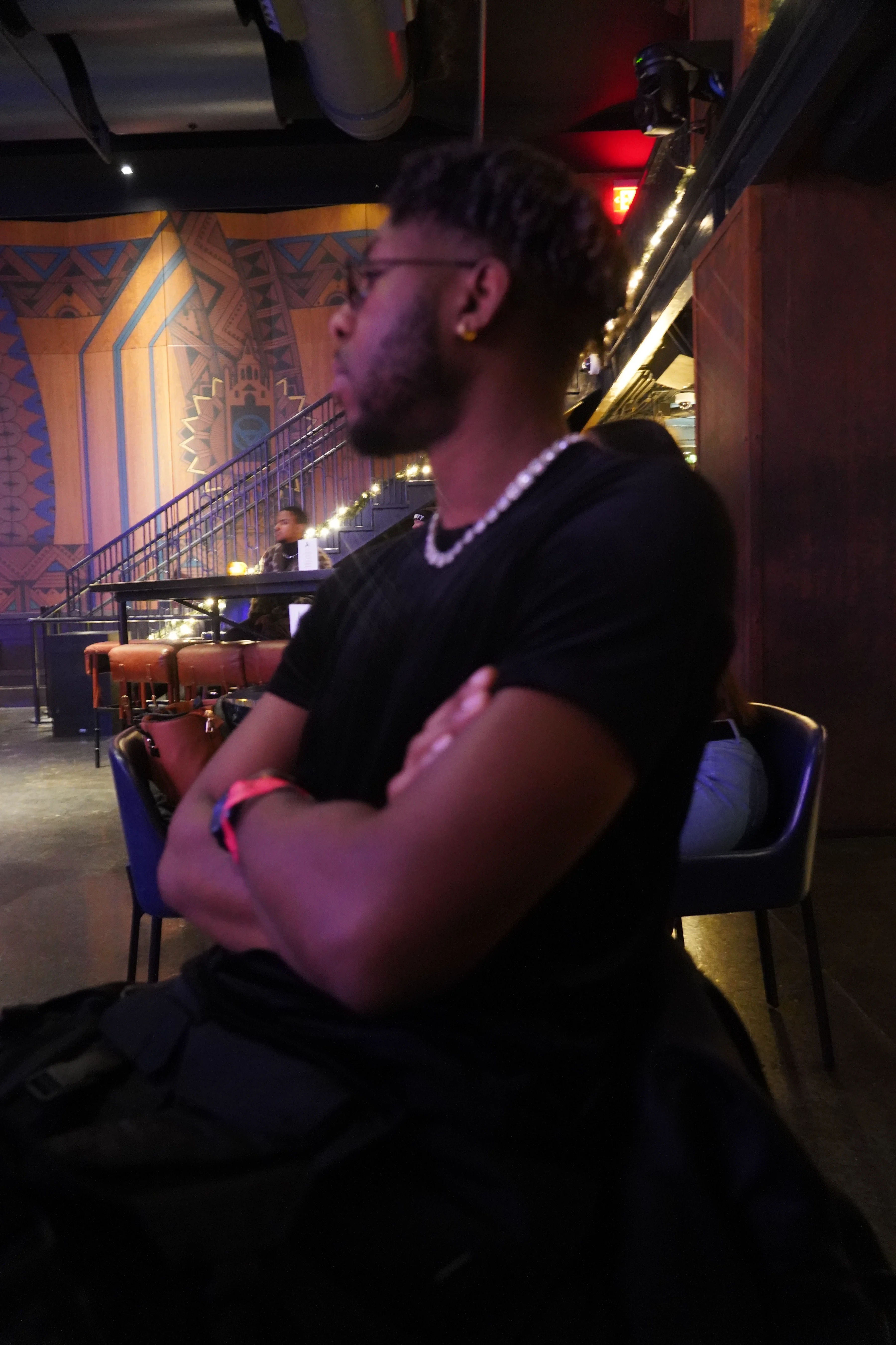 Alpha The Wolf seated in profile inside a dimly lit venue, arms crossed, gazing across the room, wearing a black crew-neck tee and pearl chain, warm amber and red light washing the ornate muraled walls behind him