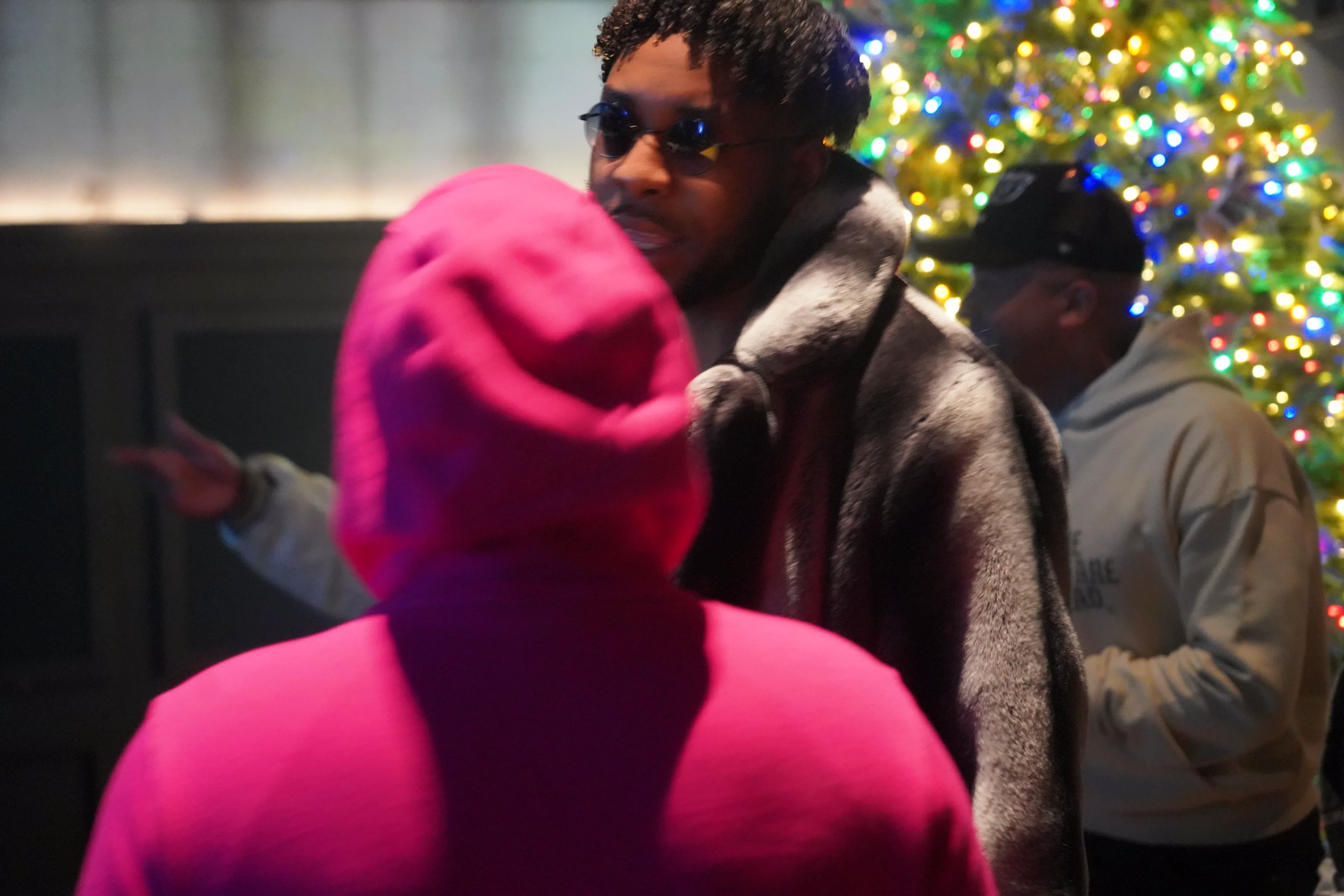 Alpha The Wolf in his grey sherpa coat, round shades and pearl chain, glancing at the camera with a measured expression while the conversation continues around him, a third guest and the Christmas tree behind him