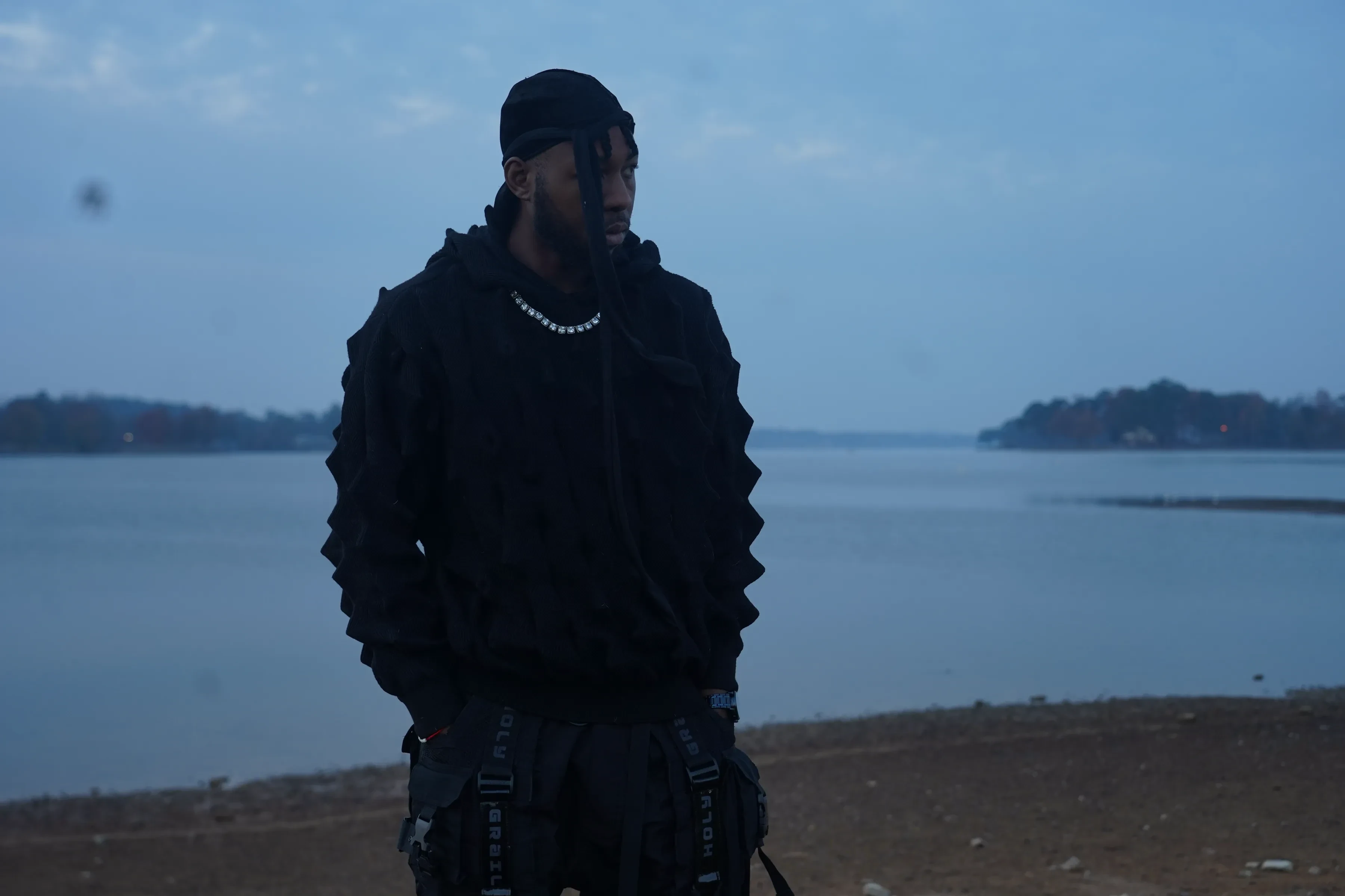 Alpha The Wolf facing the camera with hands hanging low, the ridged black hoodie holding the last blue light, a silver chain at his collar, the wide colourless lake behind him