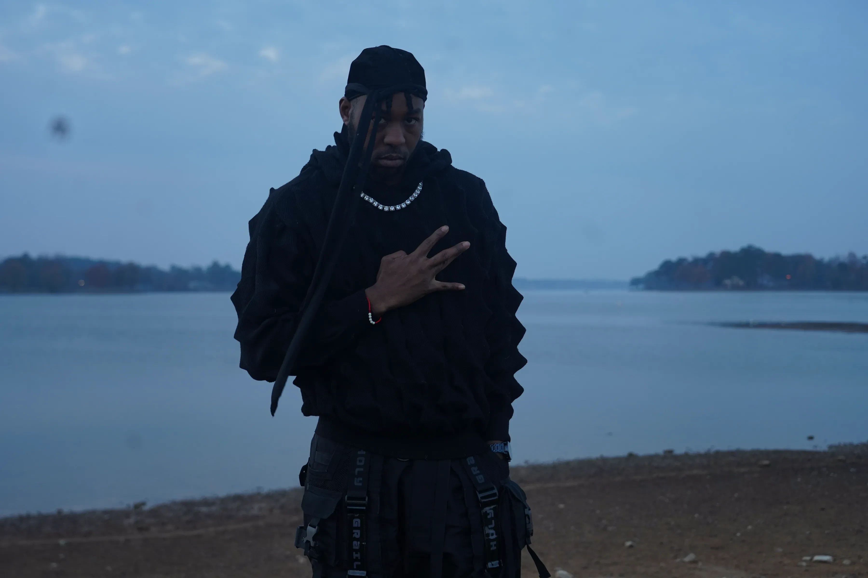 Alpha The Wolf facing the camera and throwing a hand sign at chest height, locs loose beneath the black beanie, a silver chain against the ridged hoodie, the wide empty lake behind