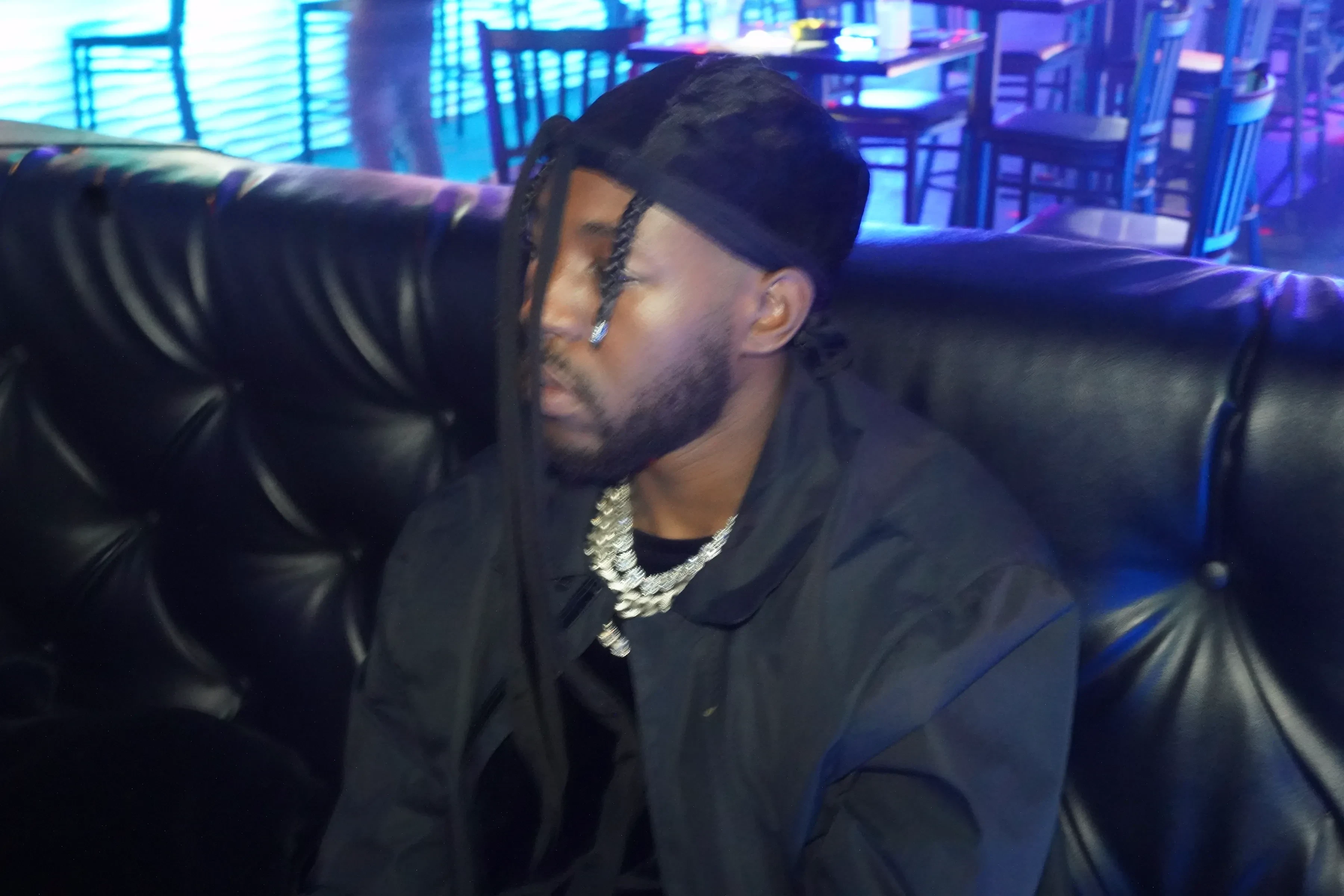 Alpha The Wolf in profile with his head slightly bowed, wearing a black velvet cap over braids with long ties trailing down, a chunky iced chain over a black tee, blue light raking across his jaw