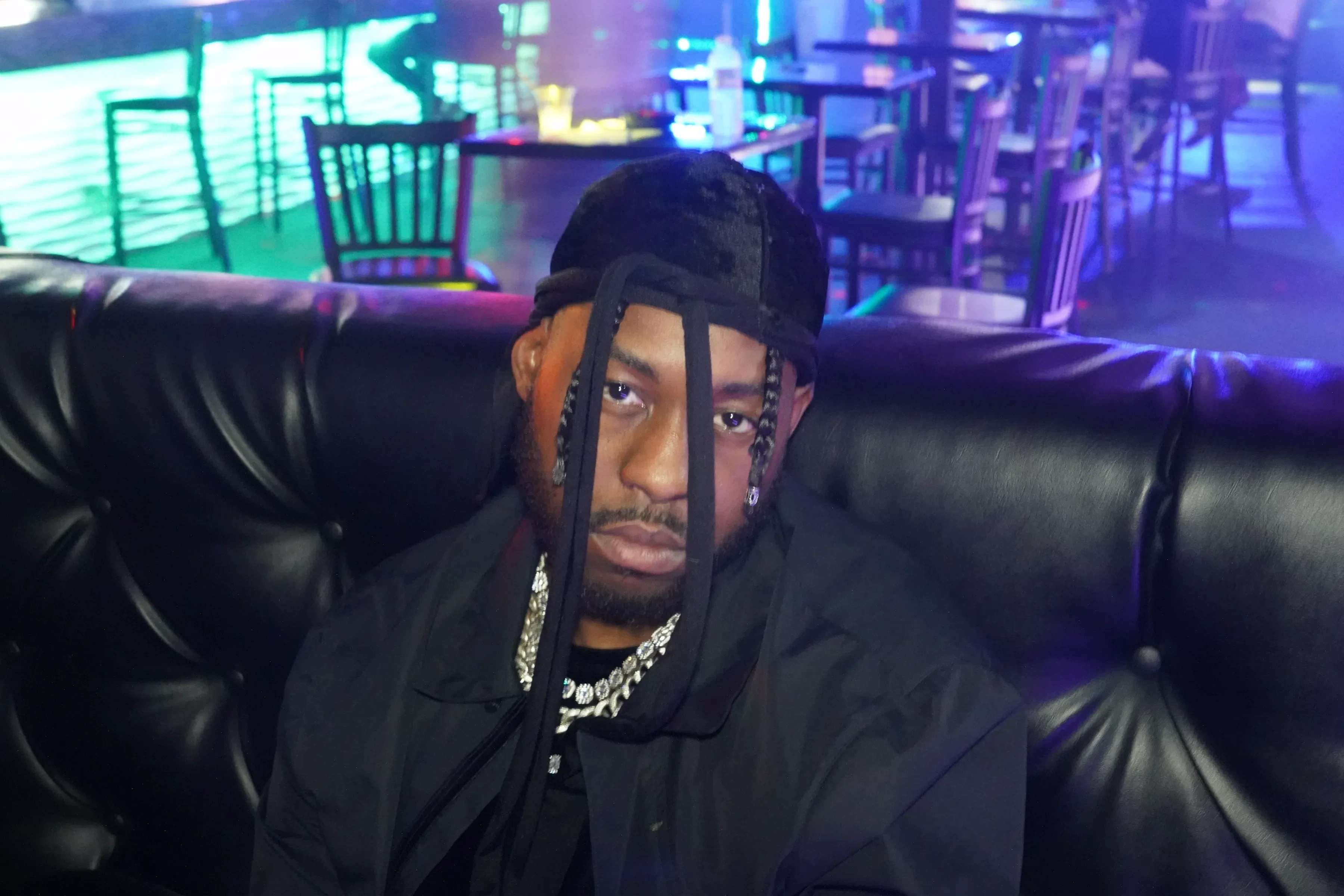 Alpha The Wolf facing the camera straight on from the booth with a flat expression, braids and durag ties framing his face, a silver chain layered over a black tee, the room lit teal and magenta