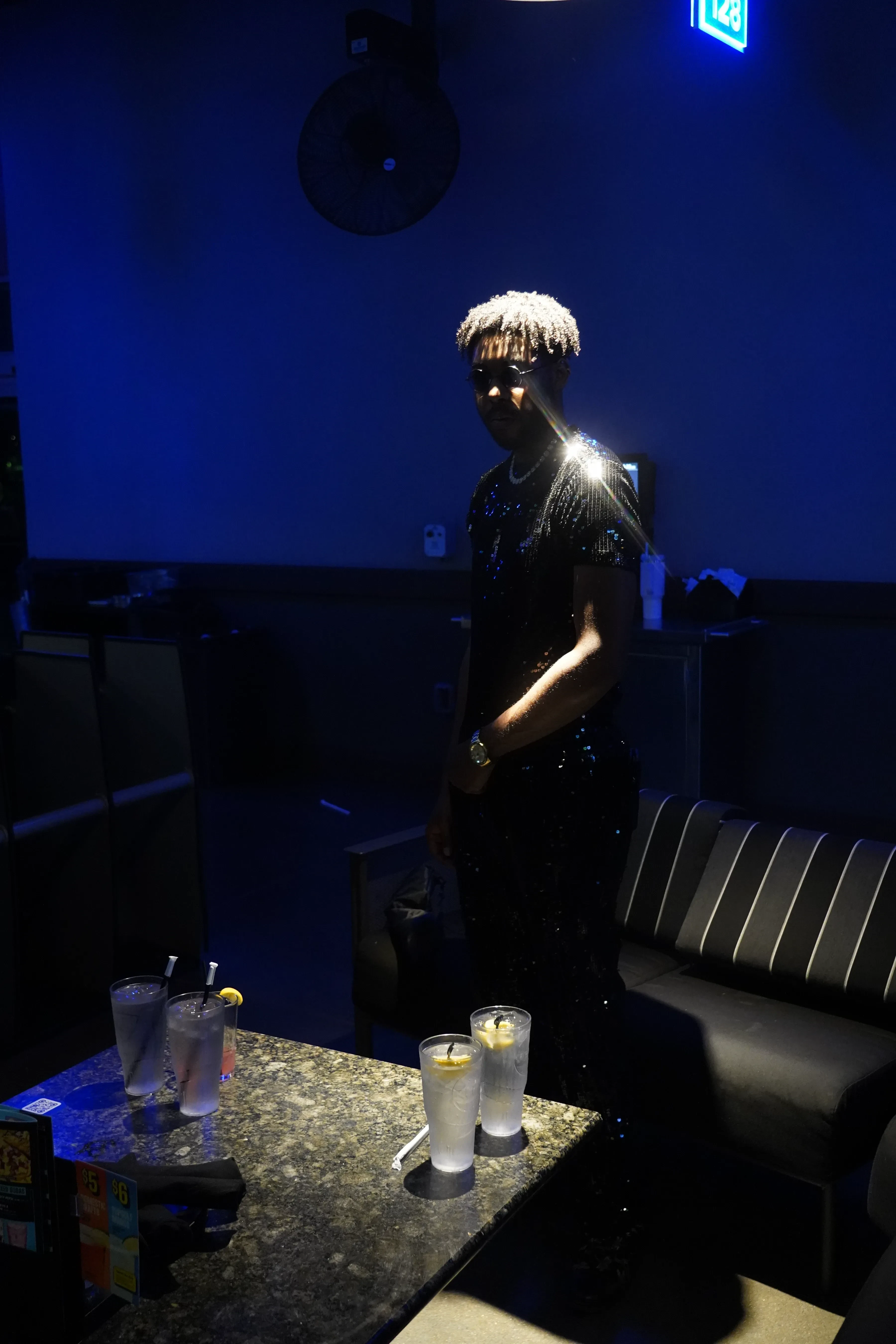 Alpha The Wolf standing in a blue-lit lounge with one hand resting on a gold watch, a narrow beam flaring off his shoulder, a granite table of iced drinks in the foreground.