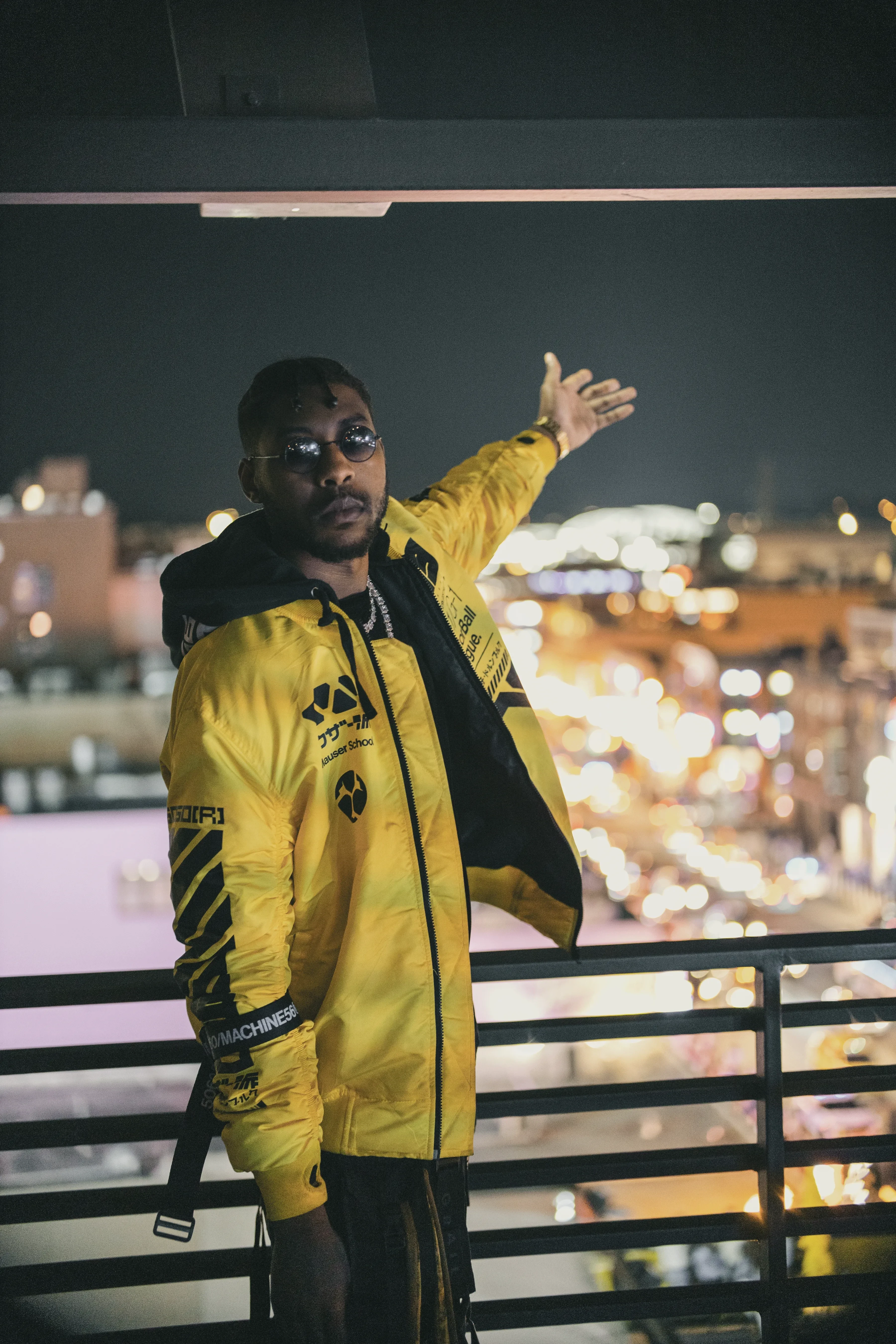 Alpha The Wolf on a balcony at night with one arm flung out to the side, the yellow bomber lit against a field of city lights blurred into bokeh behind him