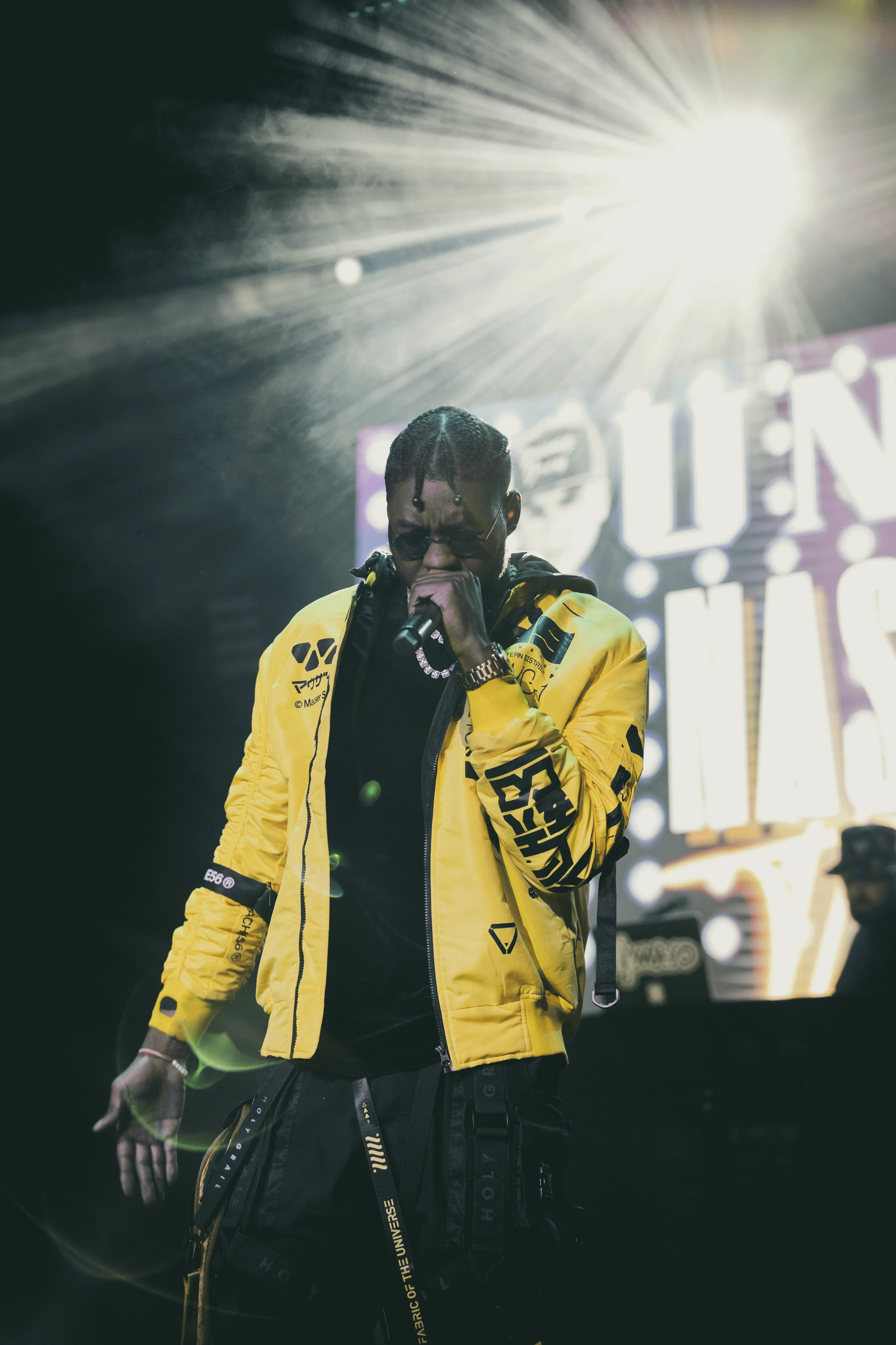Alpha The Wolf back on stage with the mic to his mouth and one hand low, a hard white beam flaring directly behind his head through the haze
